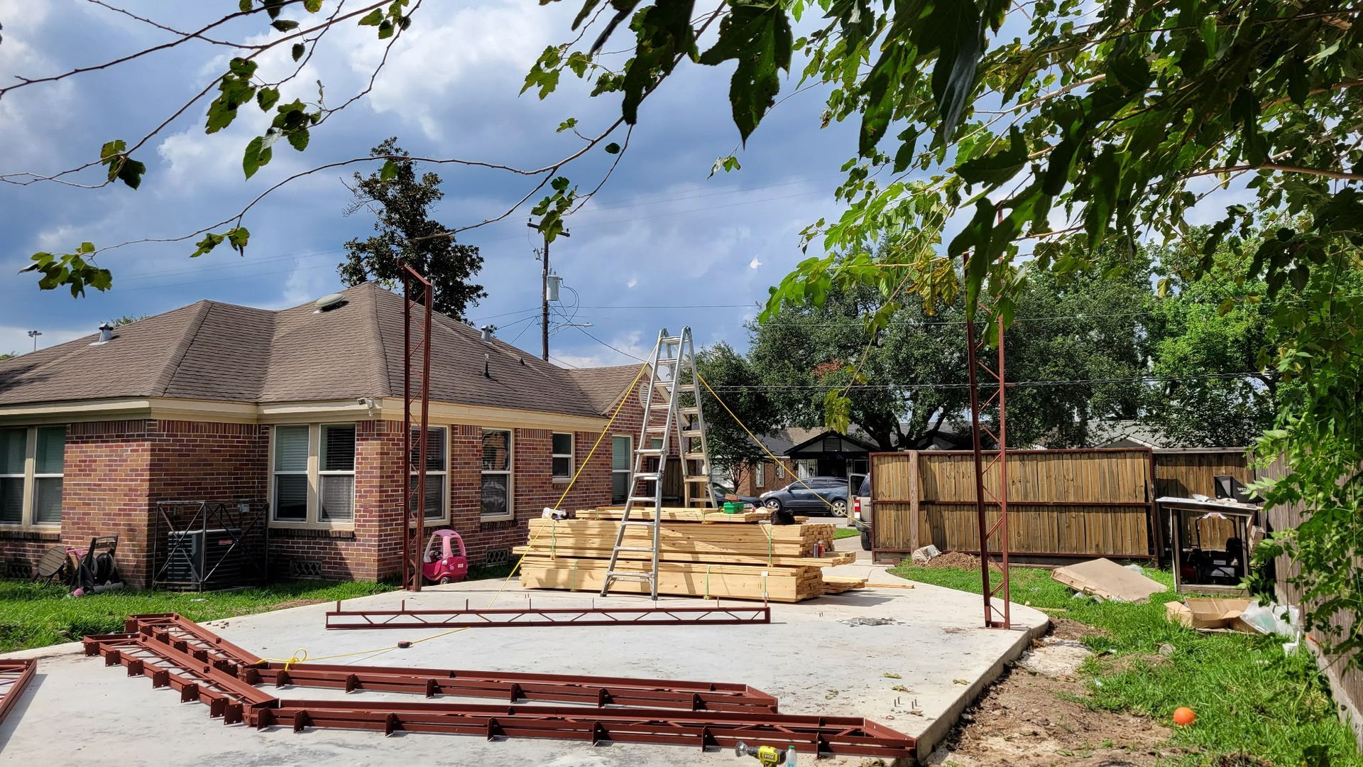Backyard construction site with a concrete slab, lumber, ladders, and a brick house.