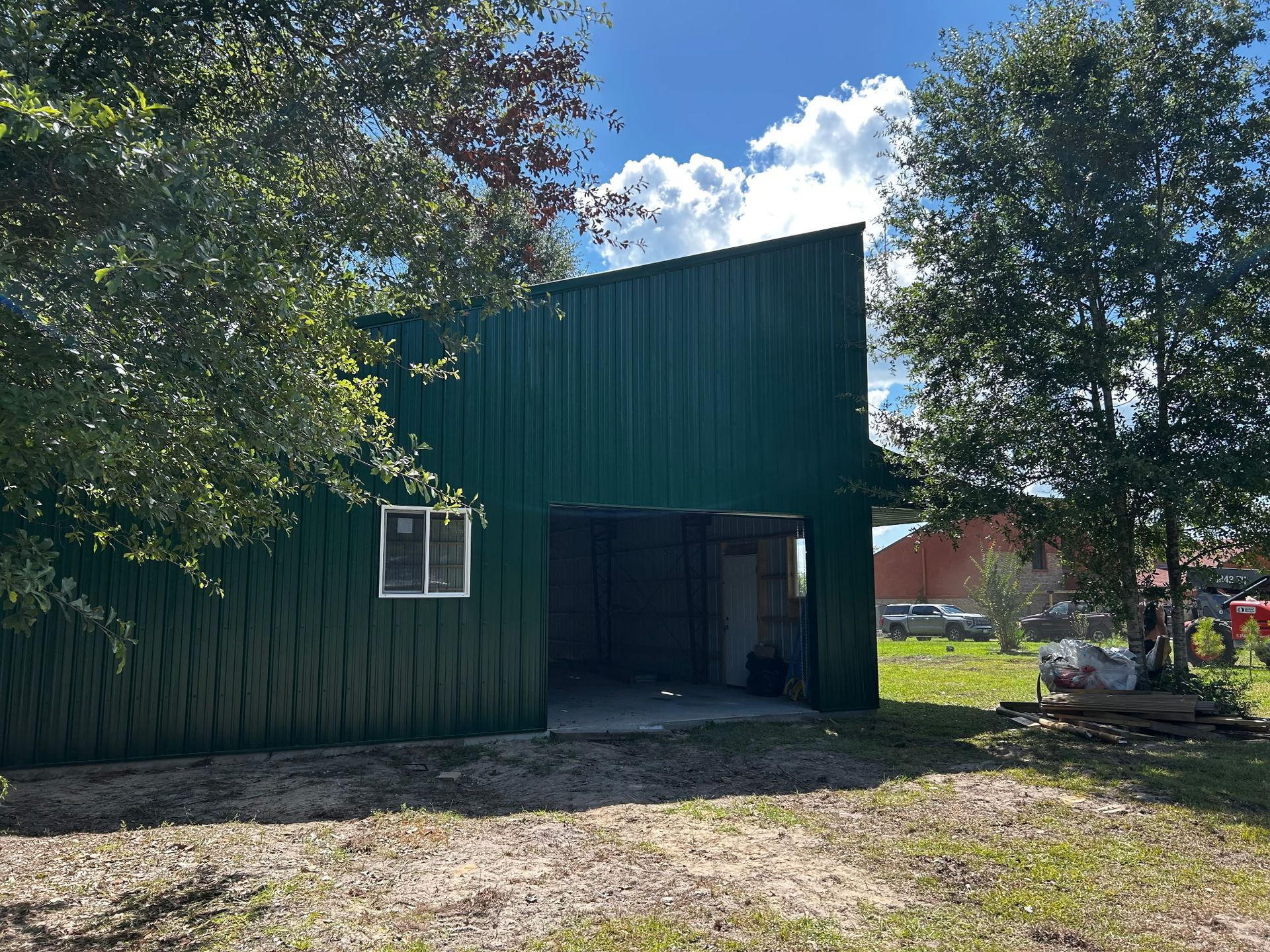 Green metal barn with open entrance under a blue sky, surrounded by trees and a few parked cars.