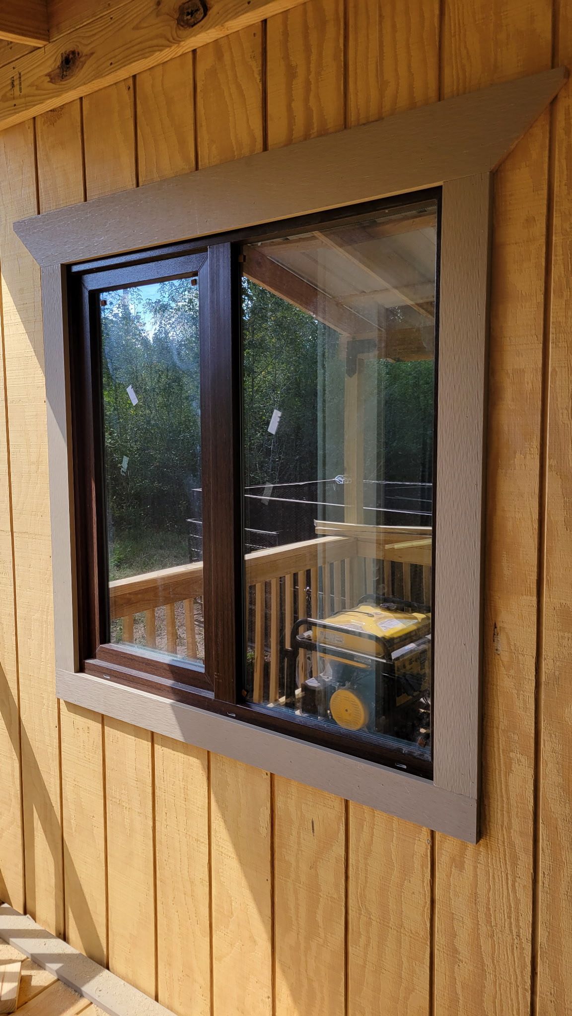 Sliding window with brown frame and gray trim set into light brown wooden siding; window reflects outdoor deck and trees.