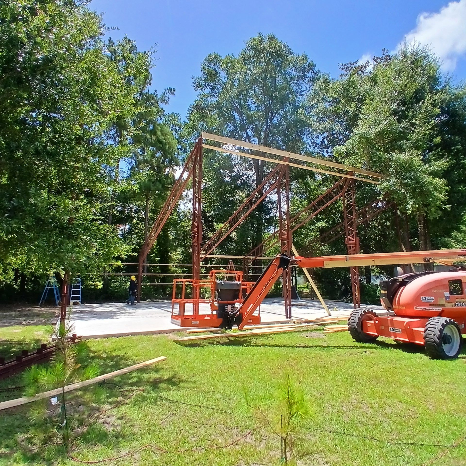 Construction of a red metal framed building with a lift in a grassy area, trees in the background.