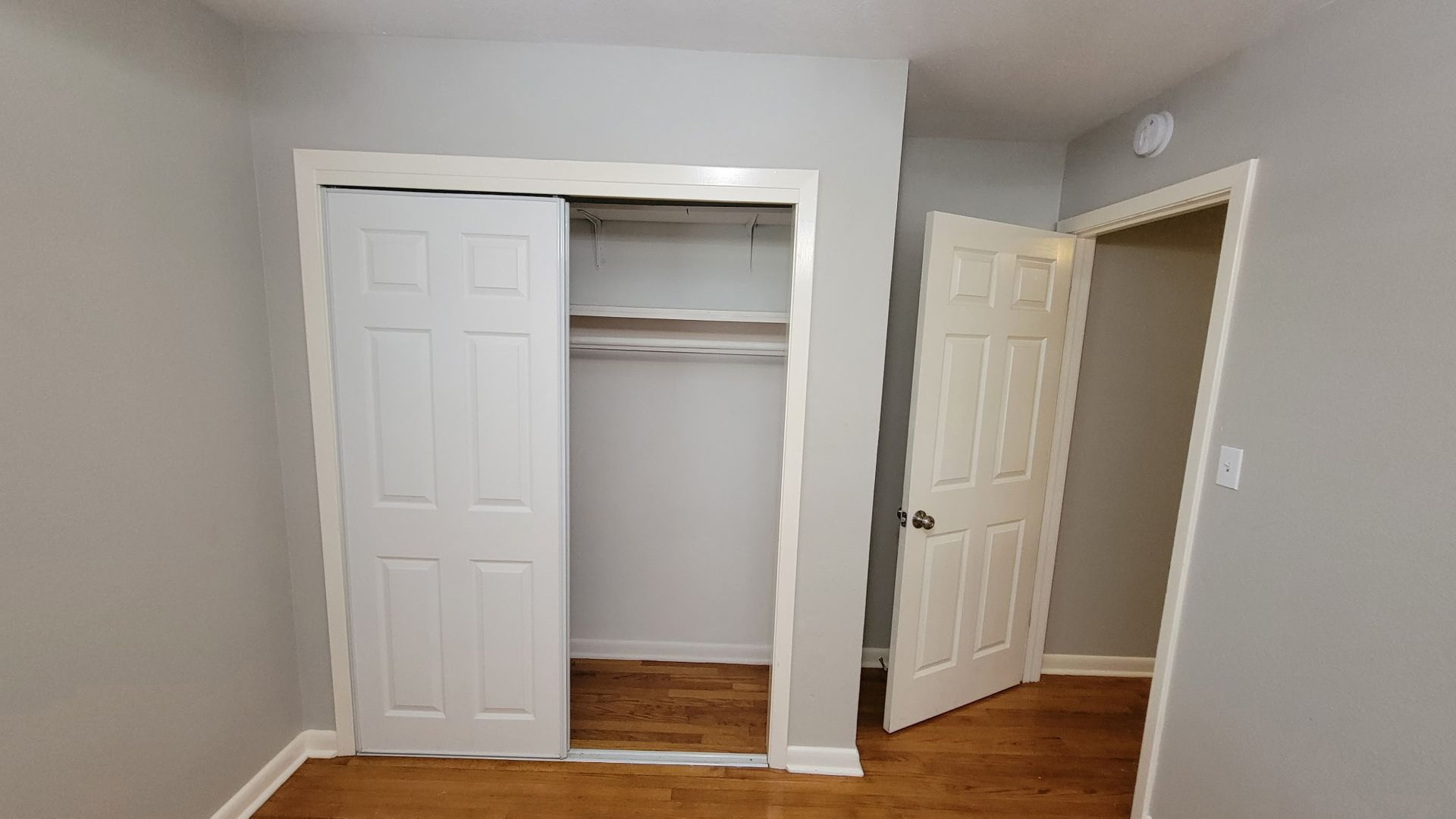 A small bedroom with an open closet and a partially open door. Light gray walls, white trim, wood floors.