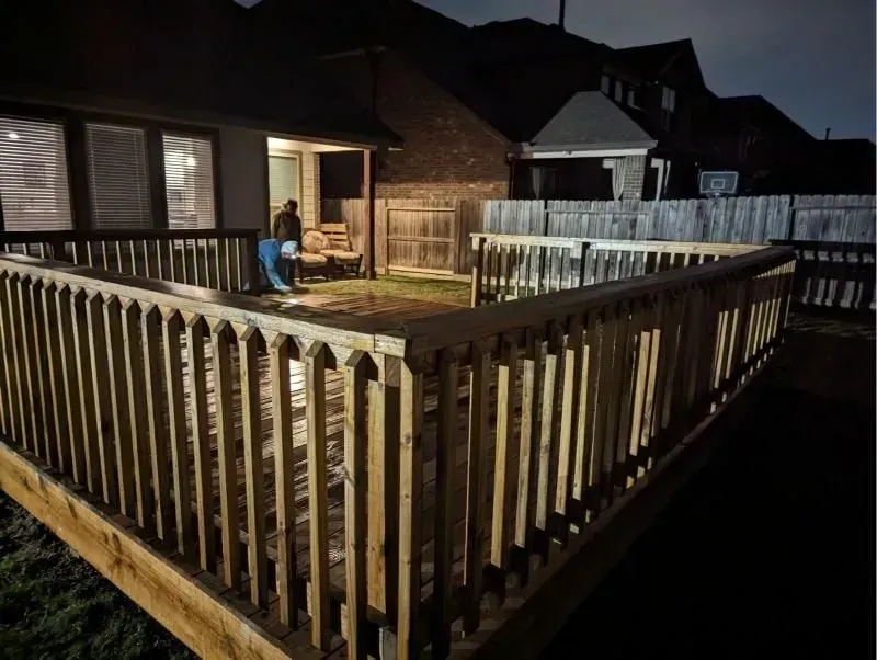 Wooden deck at night; person in the distance cleaning. Fenced backyard, lit from house, dark sky.