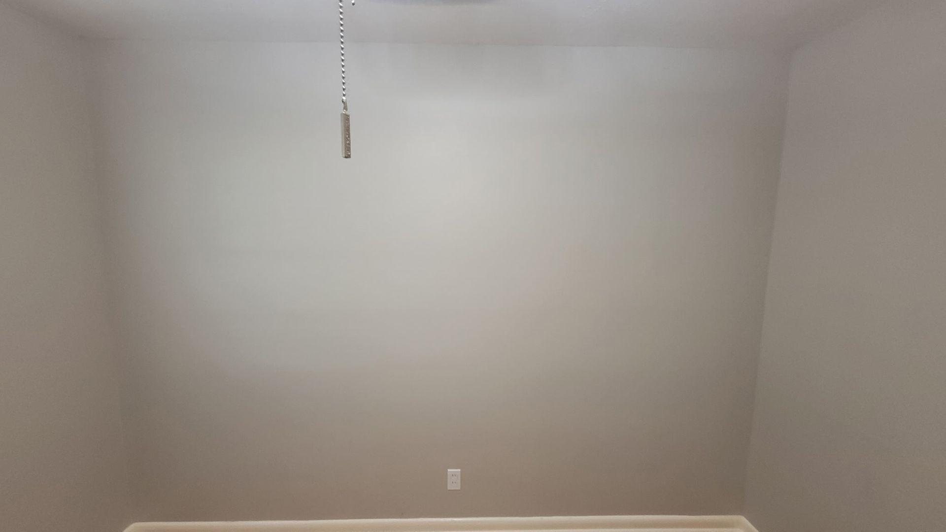 Empty room with gray walls, ceiling fan pull, and an electrical outlet.