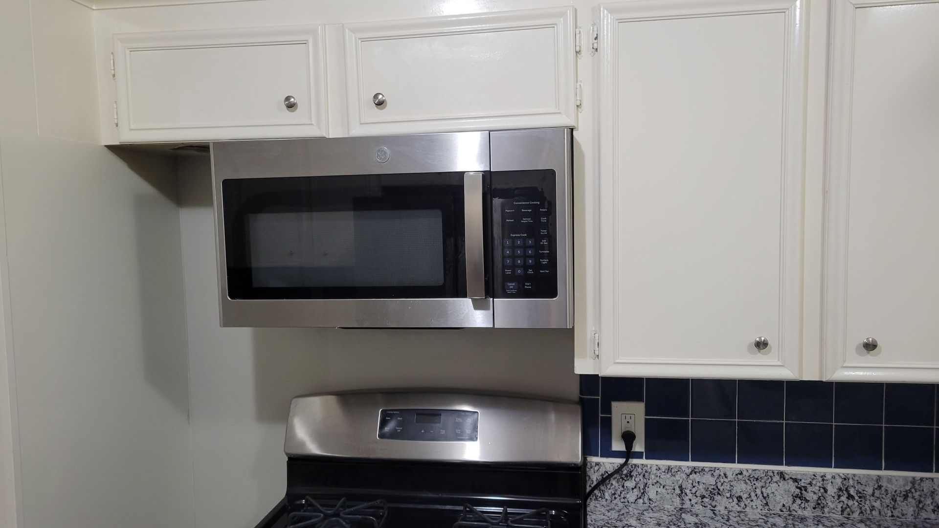 Stainless steel microwave above a stove, surrounded by white kitchen cabinets and blue tile.
