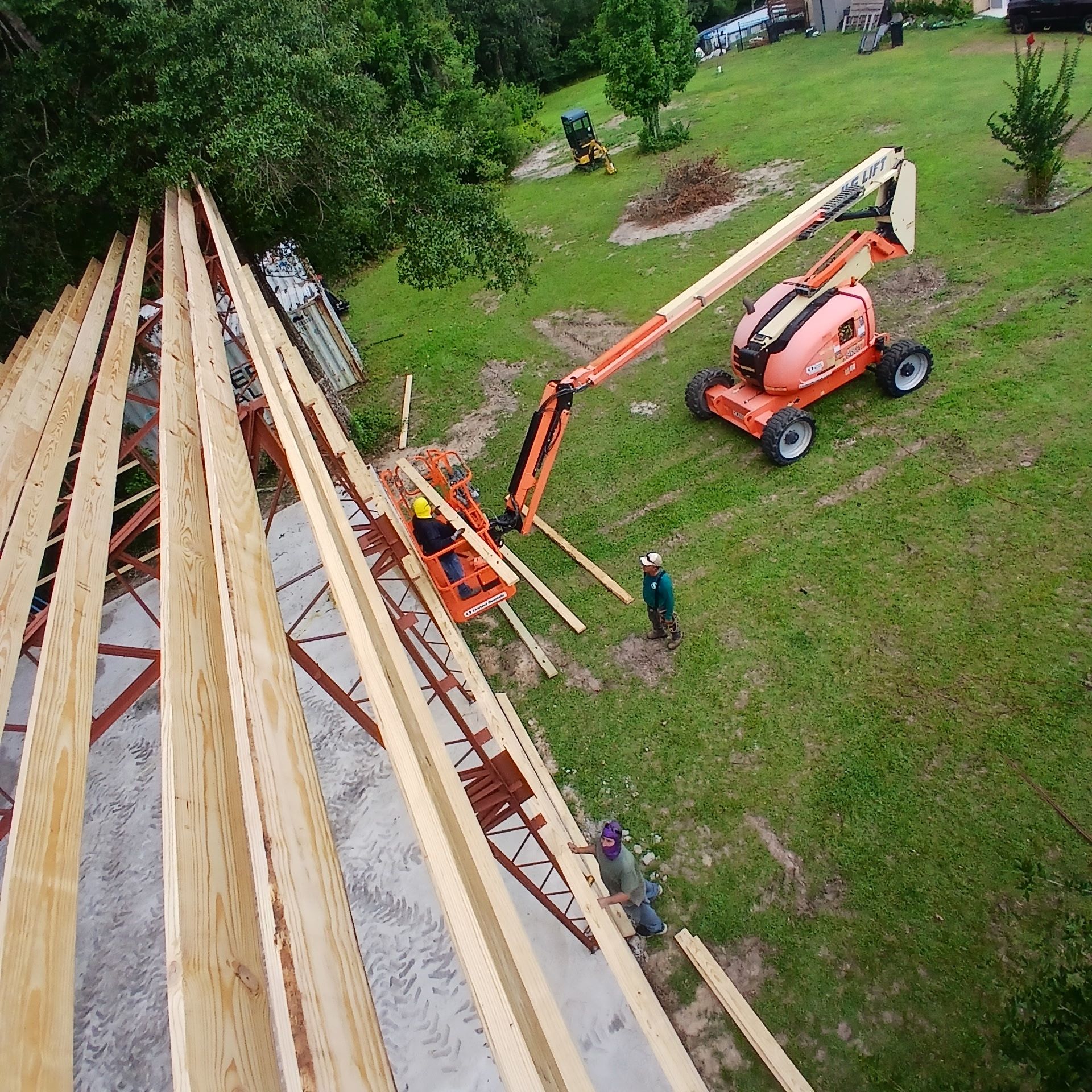 Construction workers on a roof, using a lift. Brown and orange lift, green grass, and wooden beams visible.