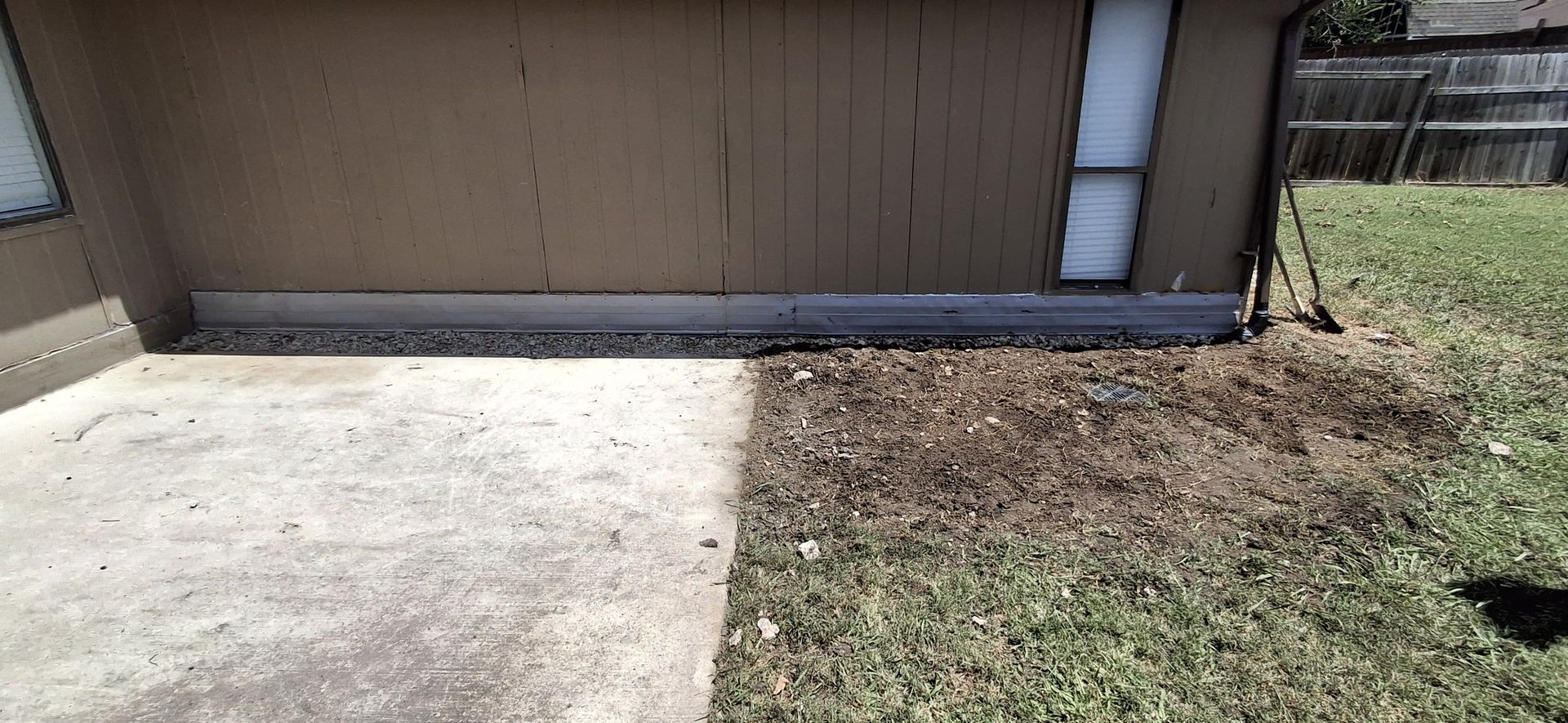 Concrete patio next to a brown building. Patchy grass and a fence are also visible.