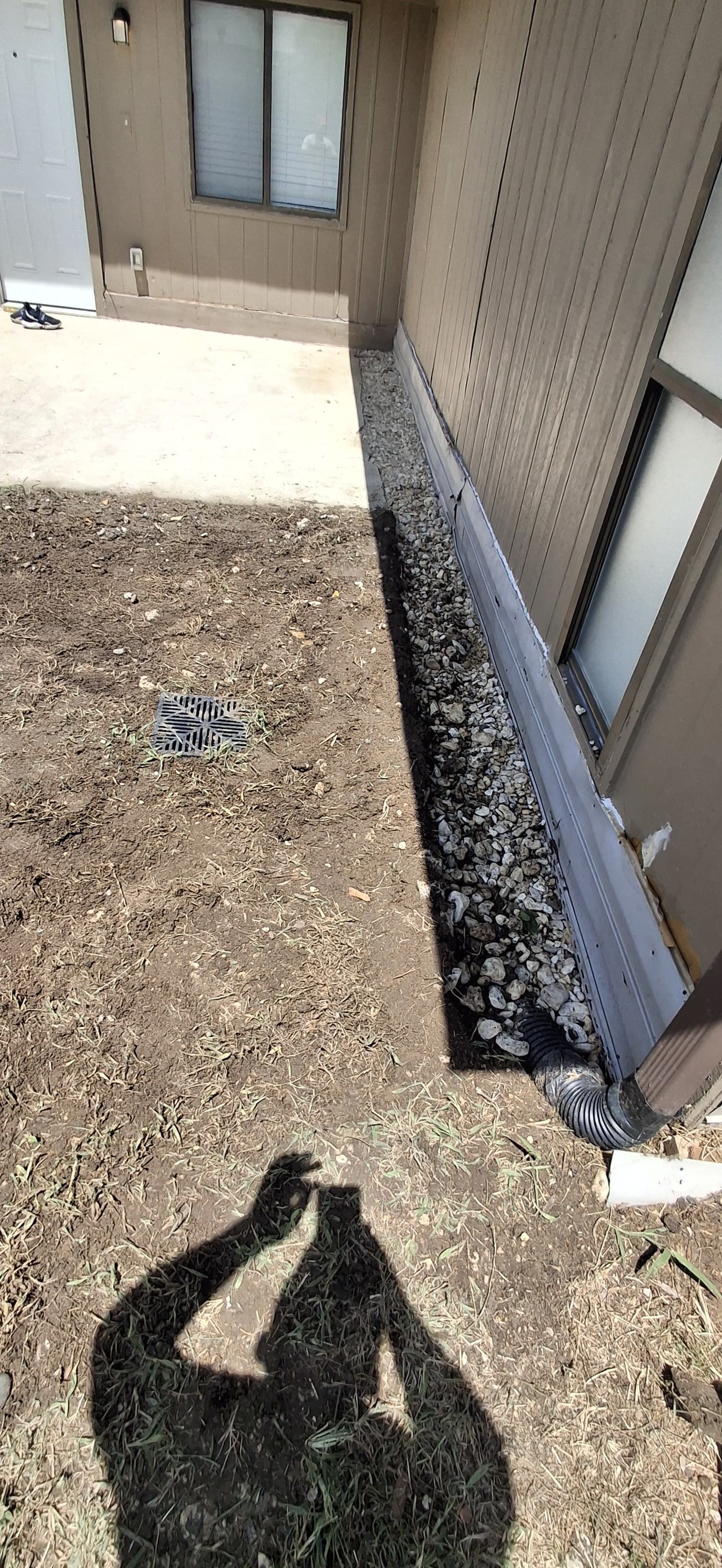 Shadow of a person taking a photo in a yard with a dirty gutter along a brick wall.