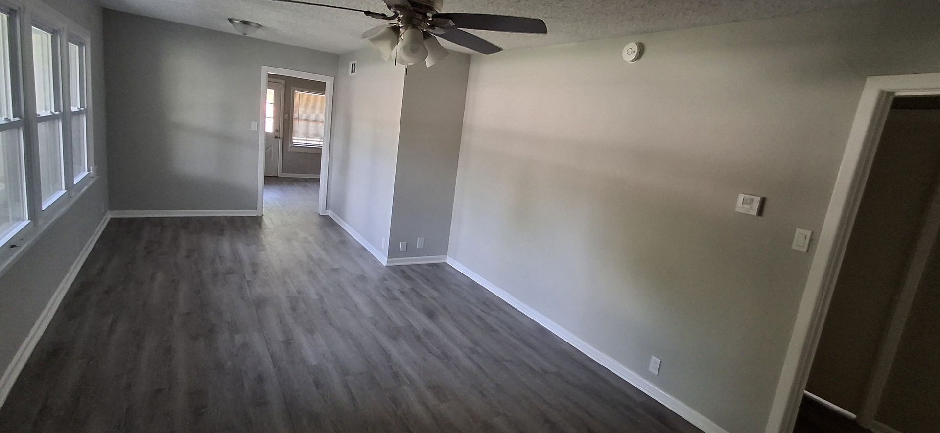 Empty living room with gray walls and flooring, a ceiling fan, and a view into a hallway.