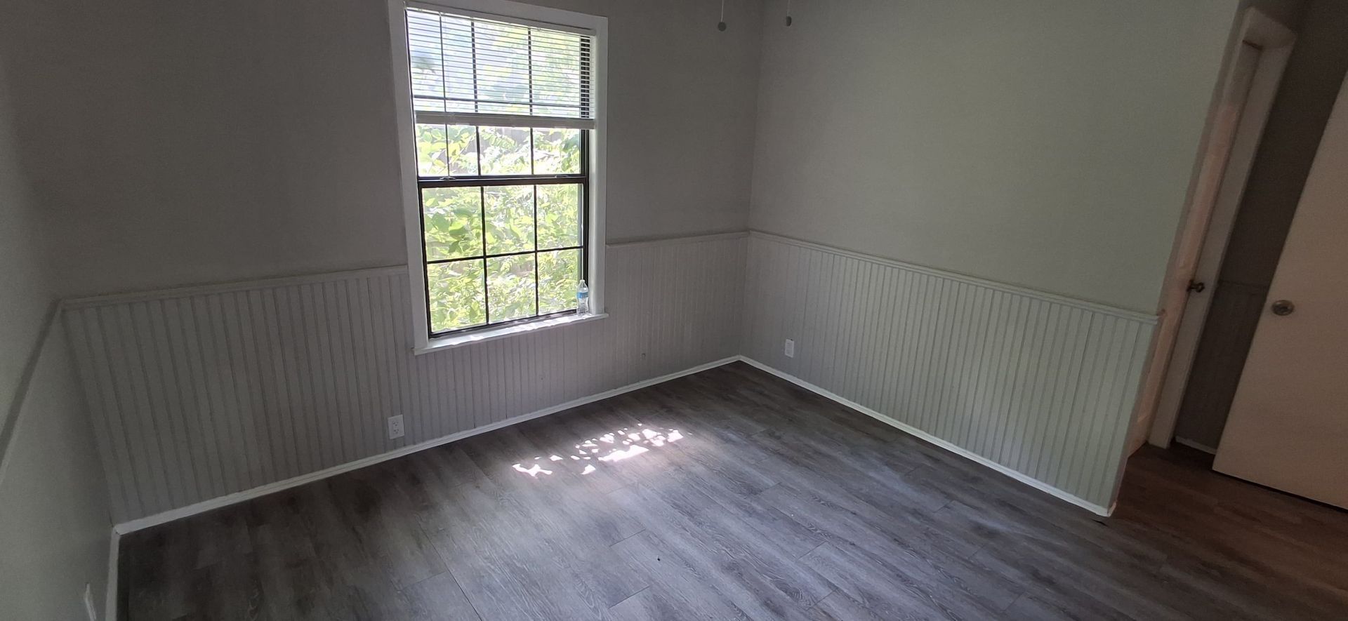 Empty room with window, wood-look flooring, light gray walls, and white paneling. A door is on the right.