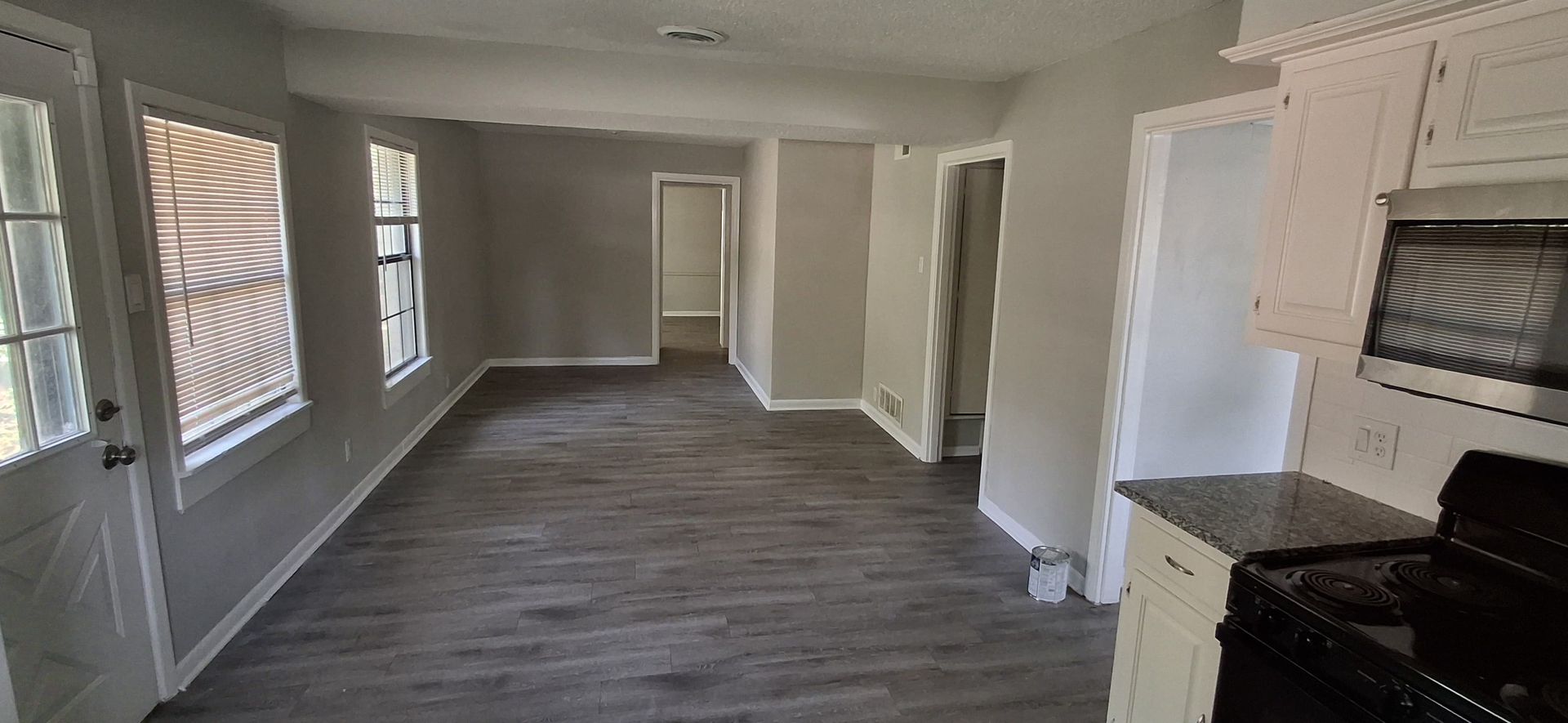 Interior view of a living space with grey walls, windows, dark wood-look flooring, and a kitchen area.