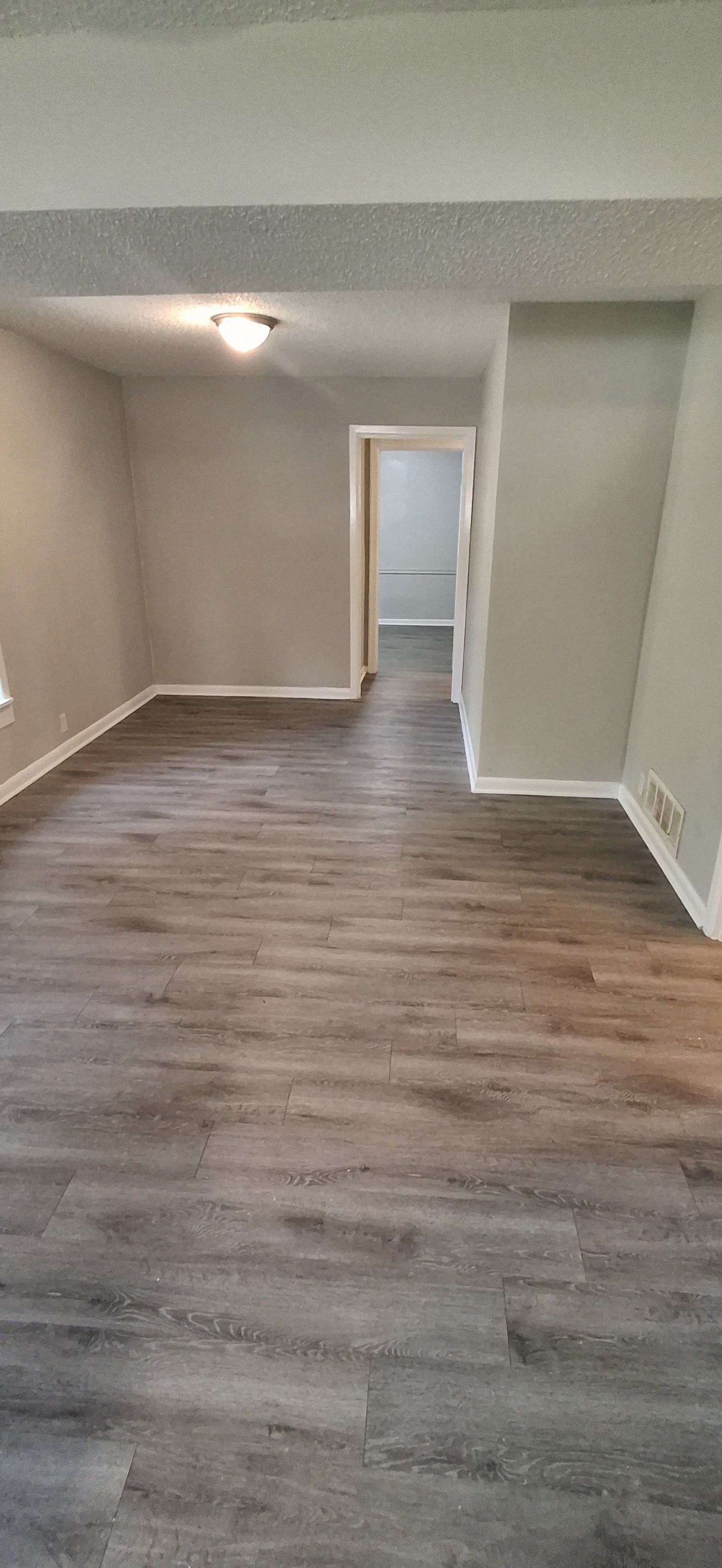 Empty room with grey walls and wood-look flooring. A hallway leads further back.