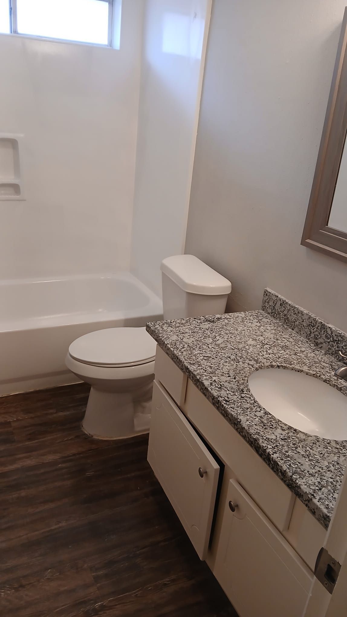 Bathroom with white tub, toilet, vanity, and brown floor. Gray countertop and wall, a window.