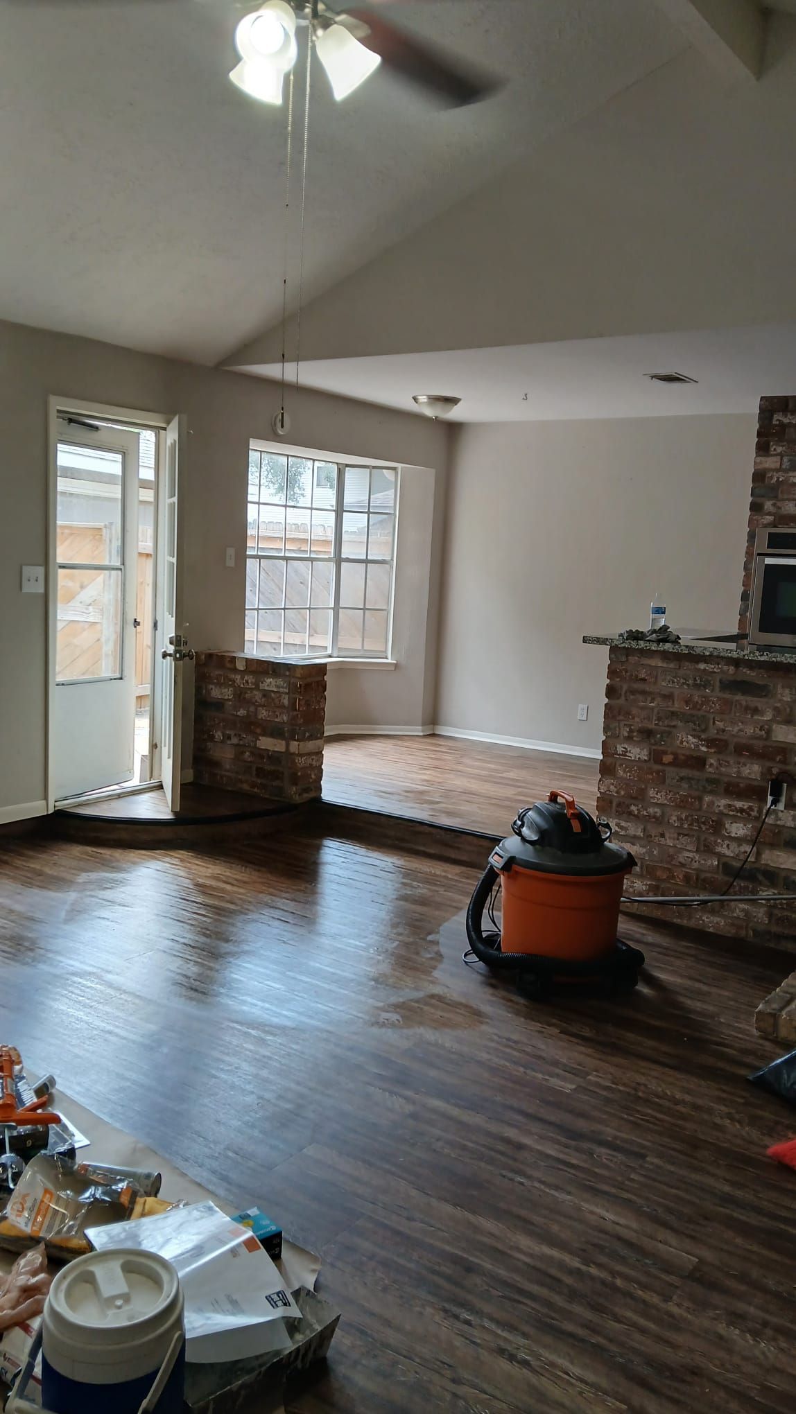 Living room with new flooring and a brick fireplace. An open doorway, a window, and a shop vacuum are present.