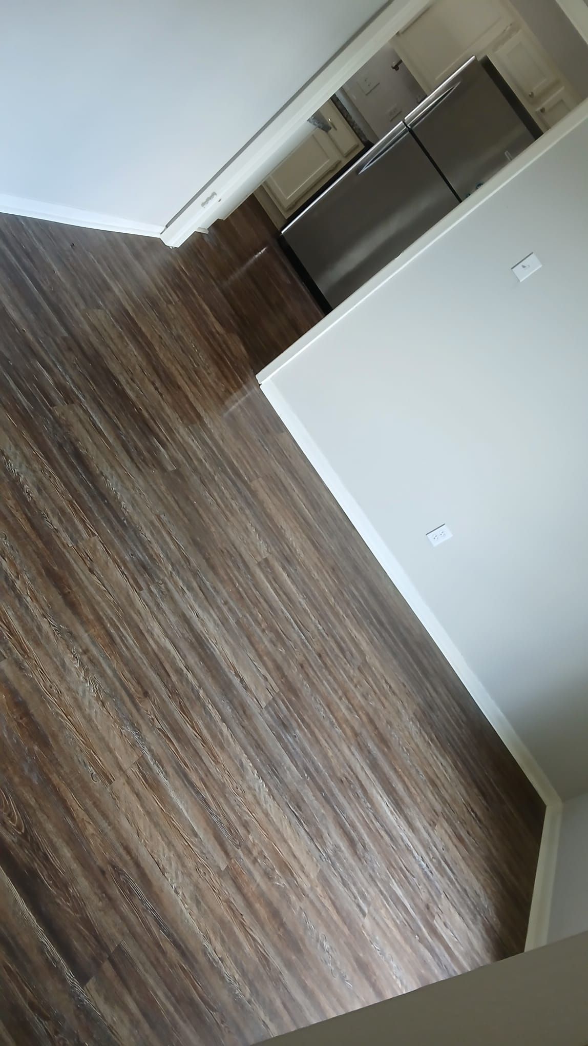Dark brown wood-look flooring leads to a kitchen area with stainless steel appliances and white walls.