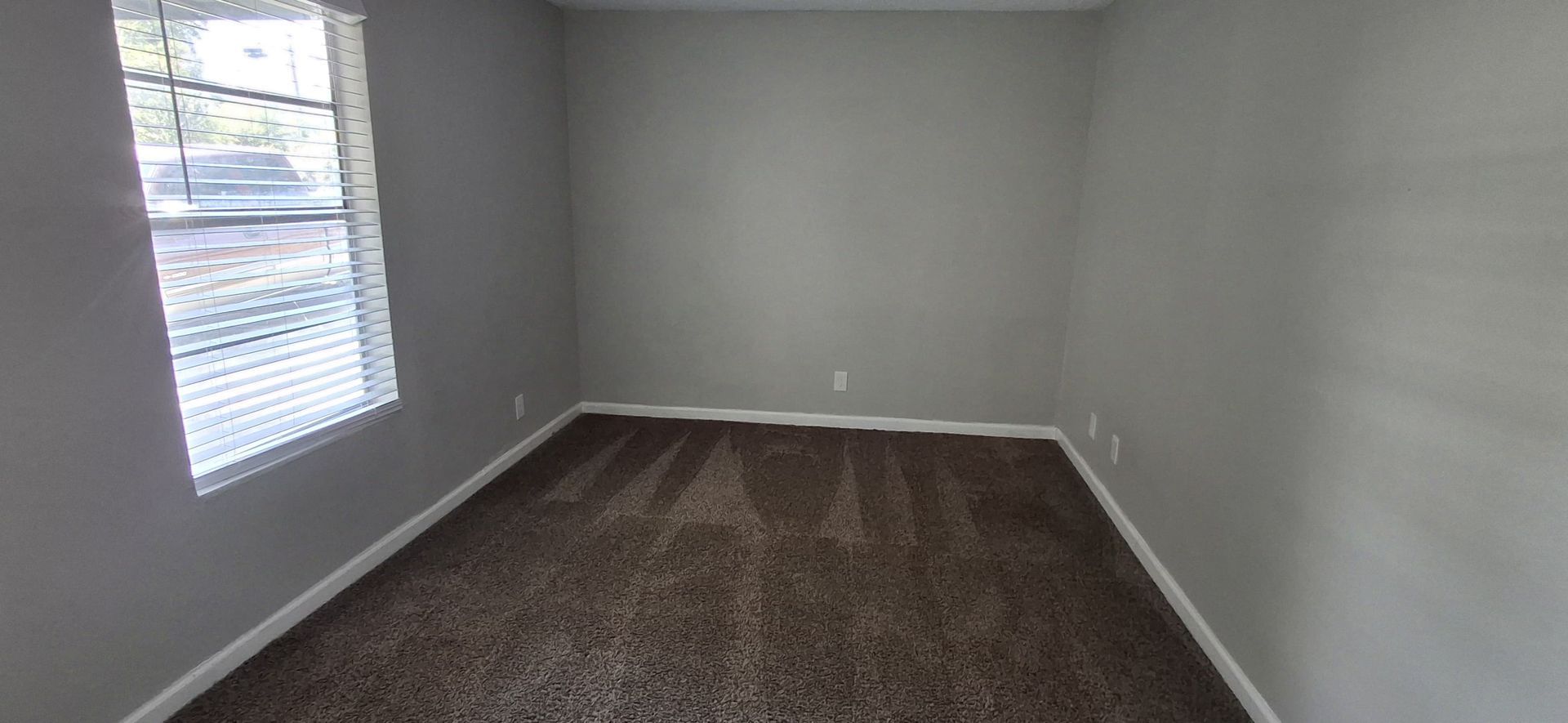 Empty room with gray walls and brown carpet; a window with blinds is on the left.