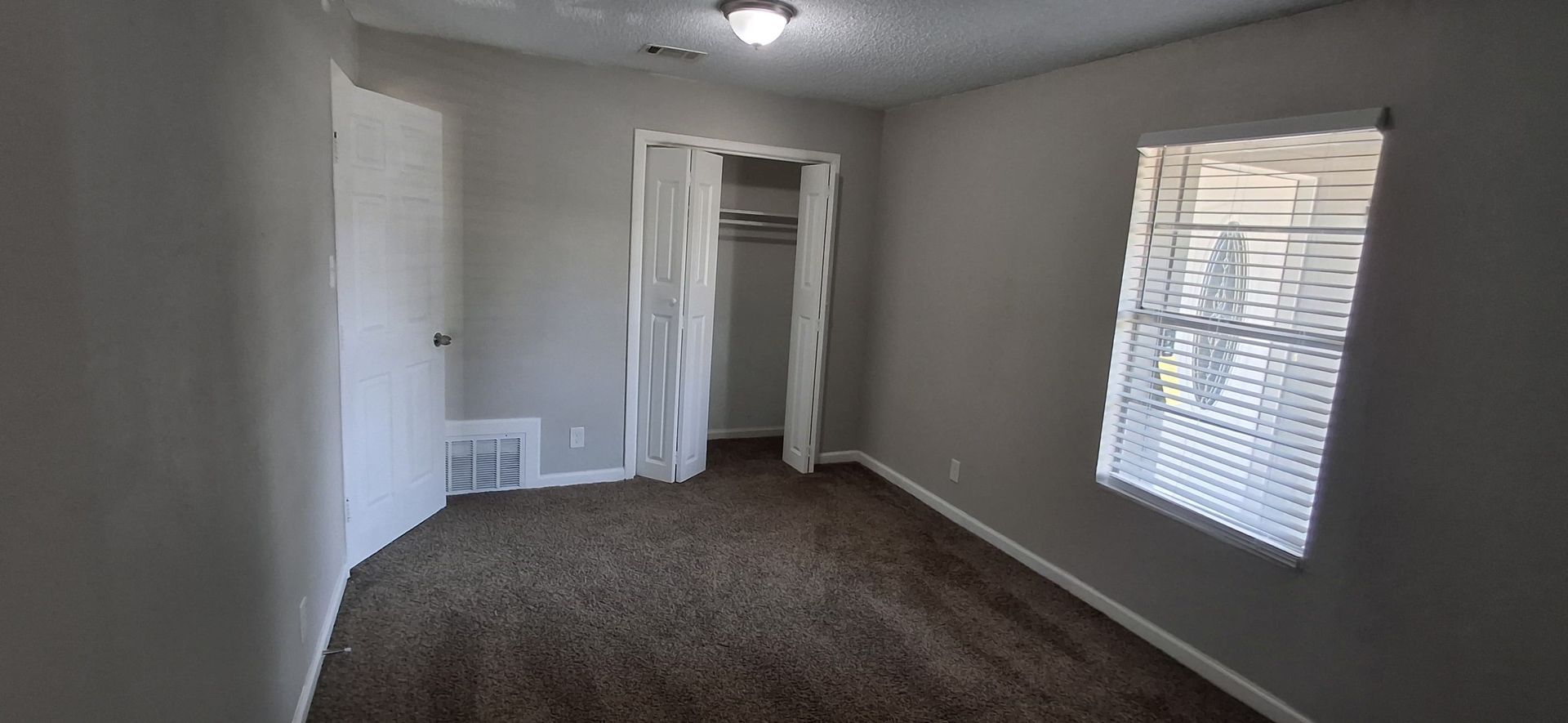 A room with grey walls, brown carpet, and a window with blinds. There is a closet with a curtain.