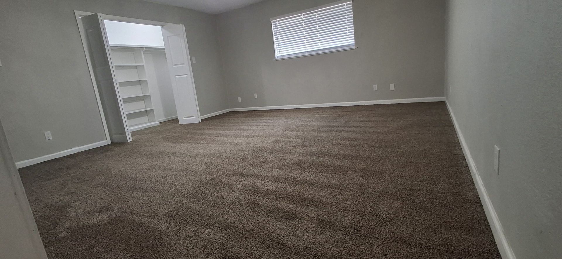Empty bedroom with brown carpet, gray walls, and a closet. Window with white curtains.