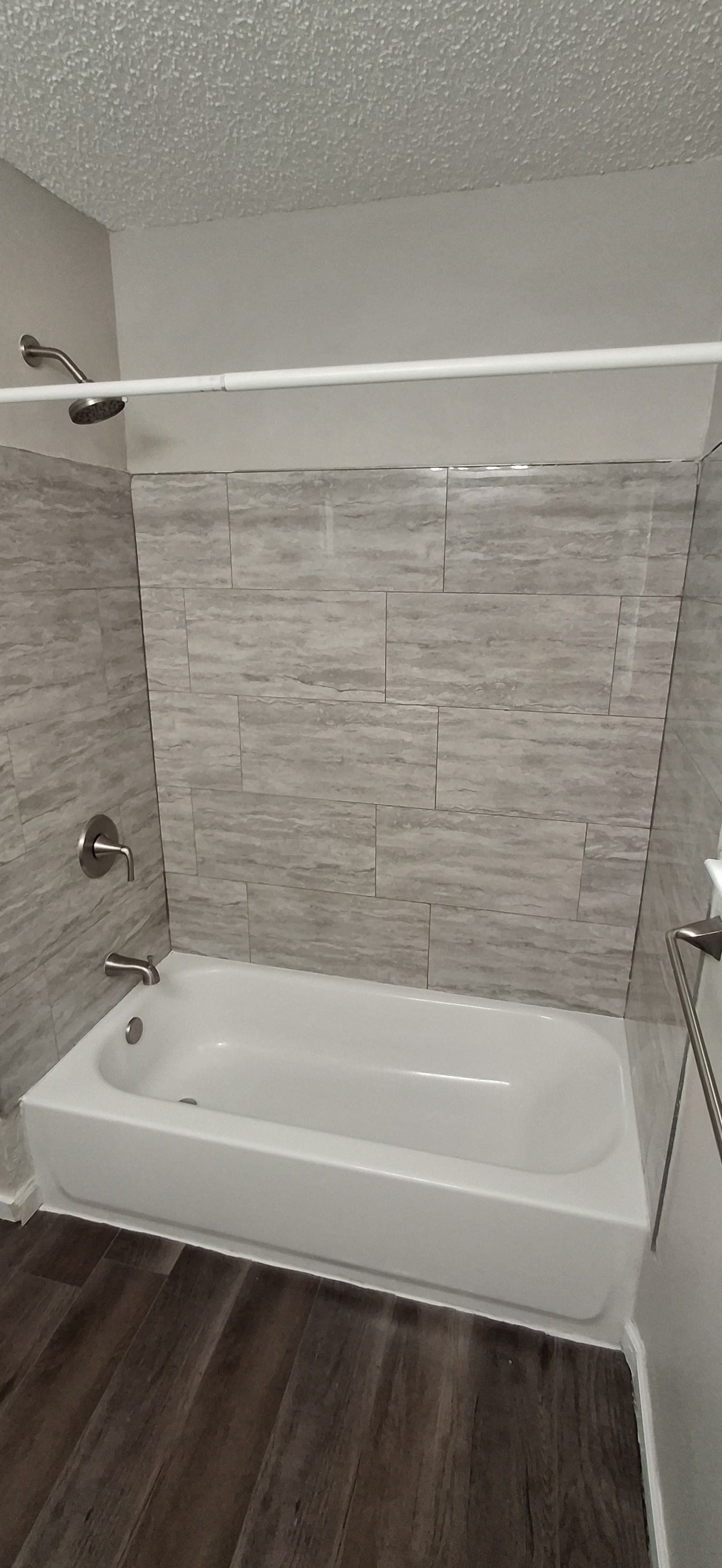 A white bathtub with gray tiled walls and a dark wood floor. Chrome shower fixtures.