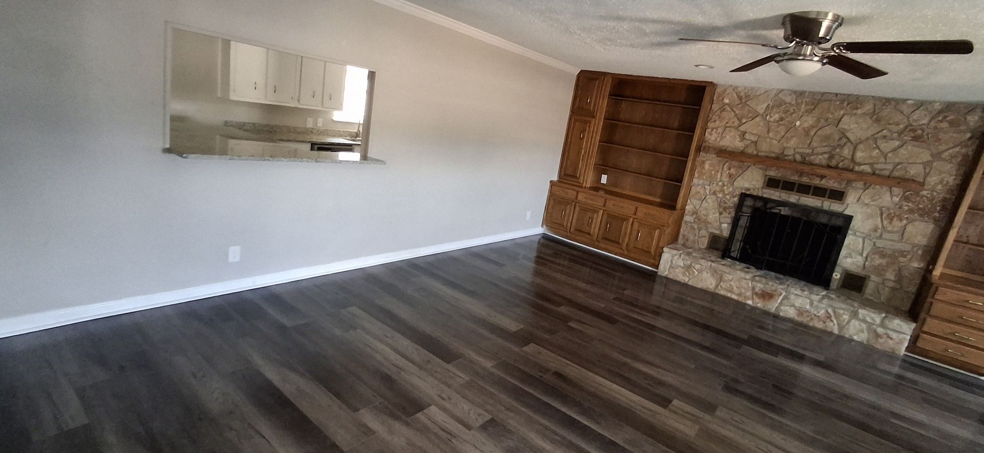 Living room with a stone fireplace, wood shelving, and dark wood-look flooring. A mirror reflects a kitchen.