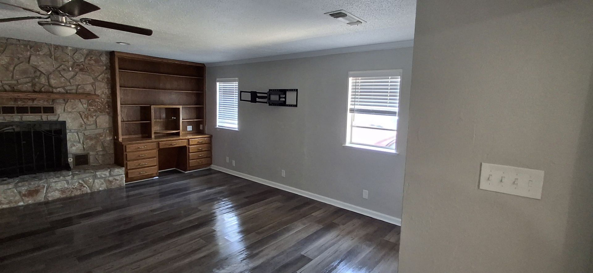 Living room with stone fireplace, wooden desk, grey walls, two windows, and dark wood flooring.