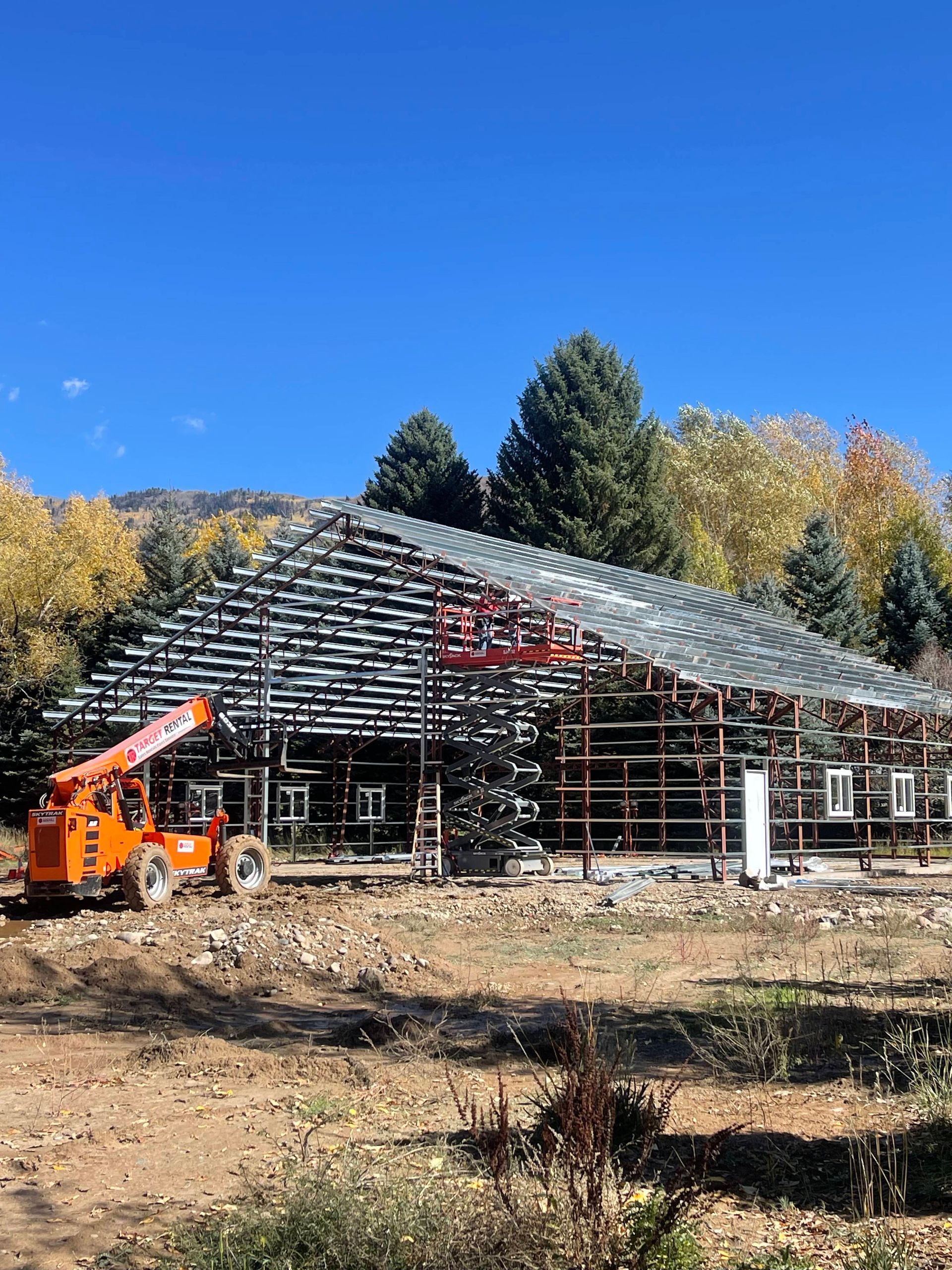 Construction site of a building with visible framework; orange lift crane. Blue sky, fall foliage in the background.