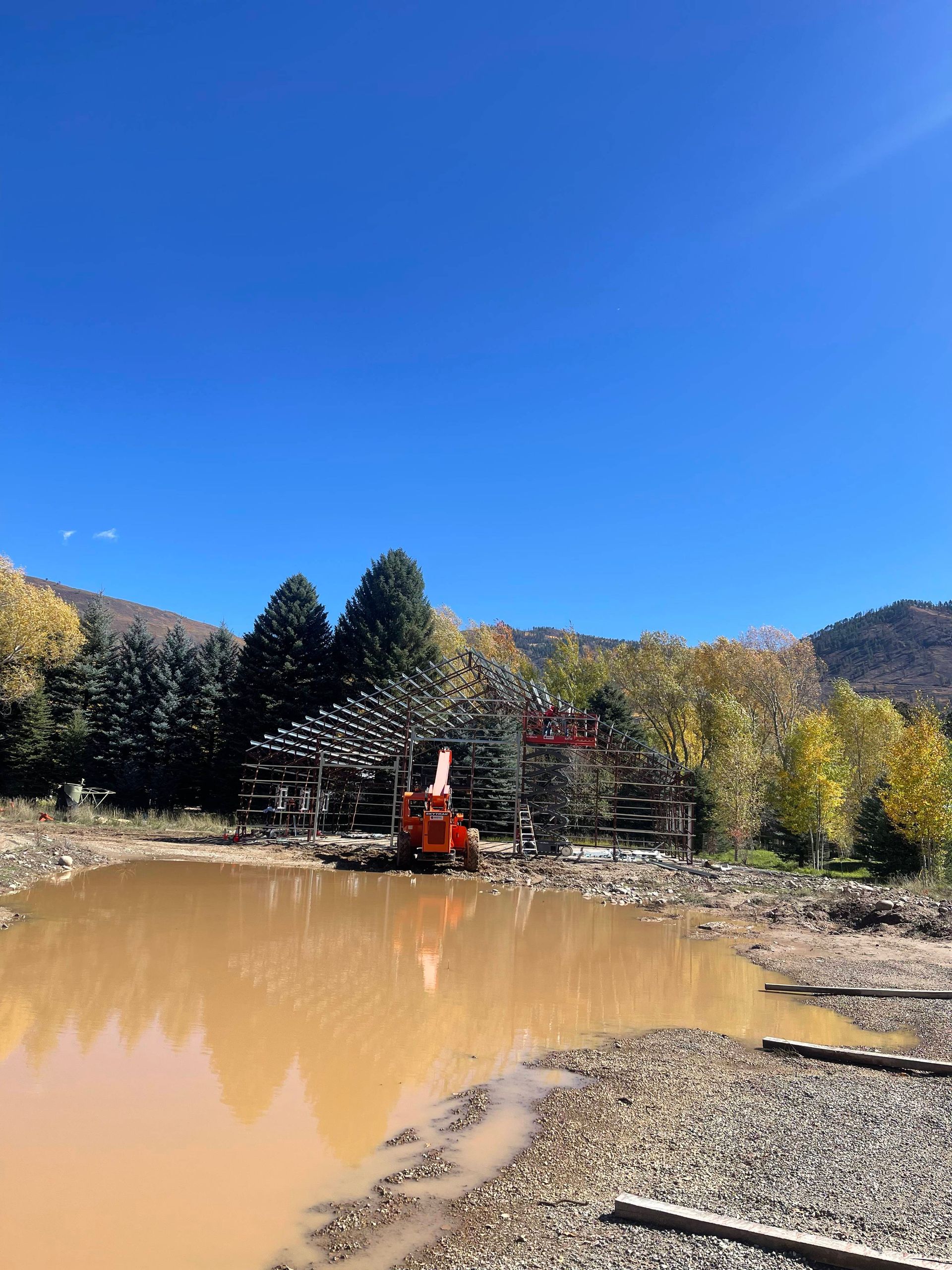 Orange excavator works in muddy water with surrounding trees under blue sky.