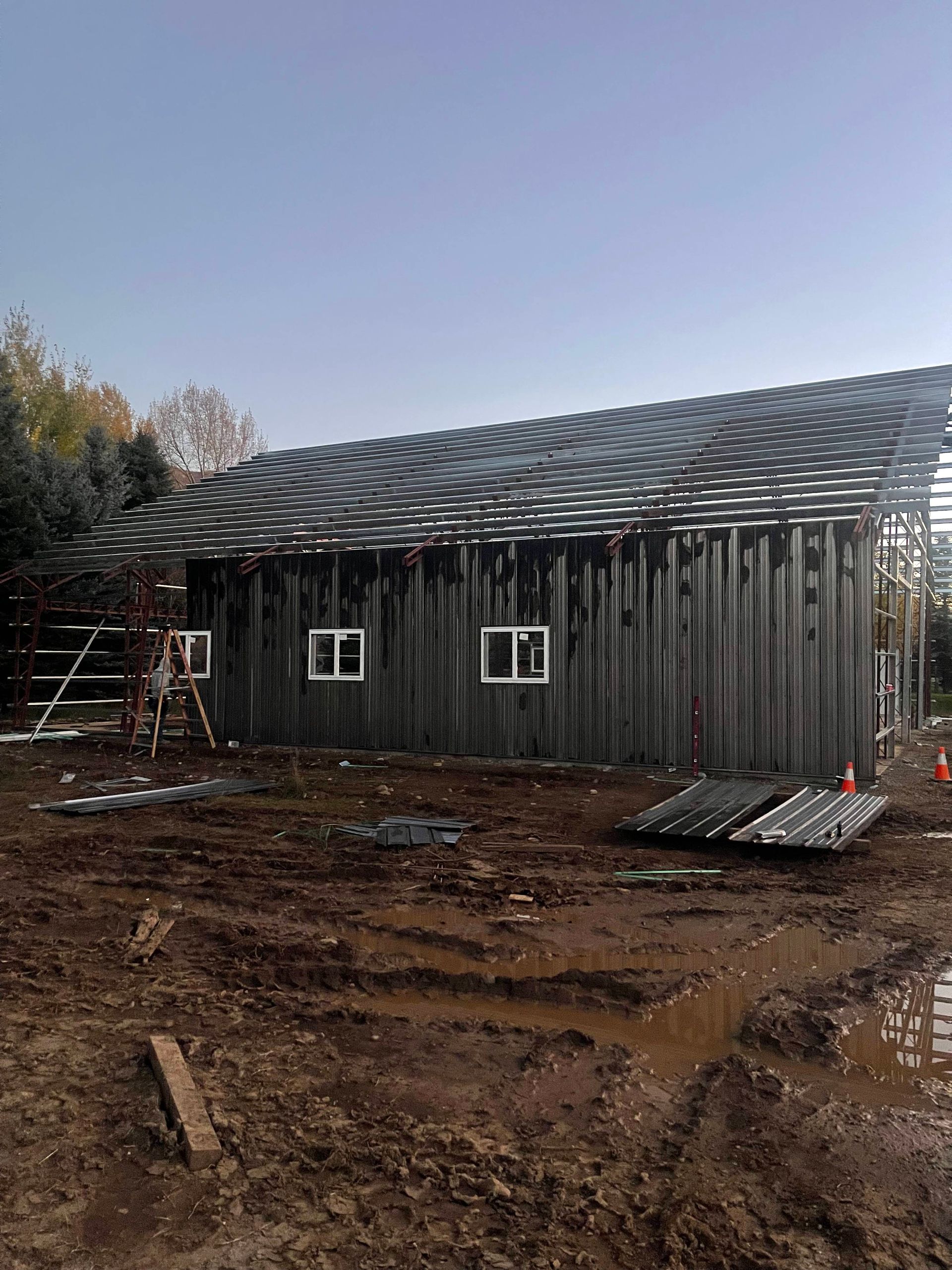 Construction site: partially built gray building with corrugated metal roof under a blue sky, surrounded by mud.