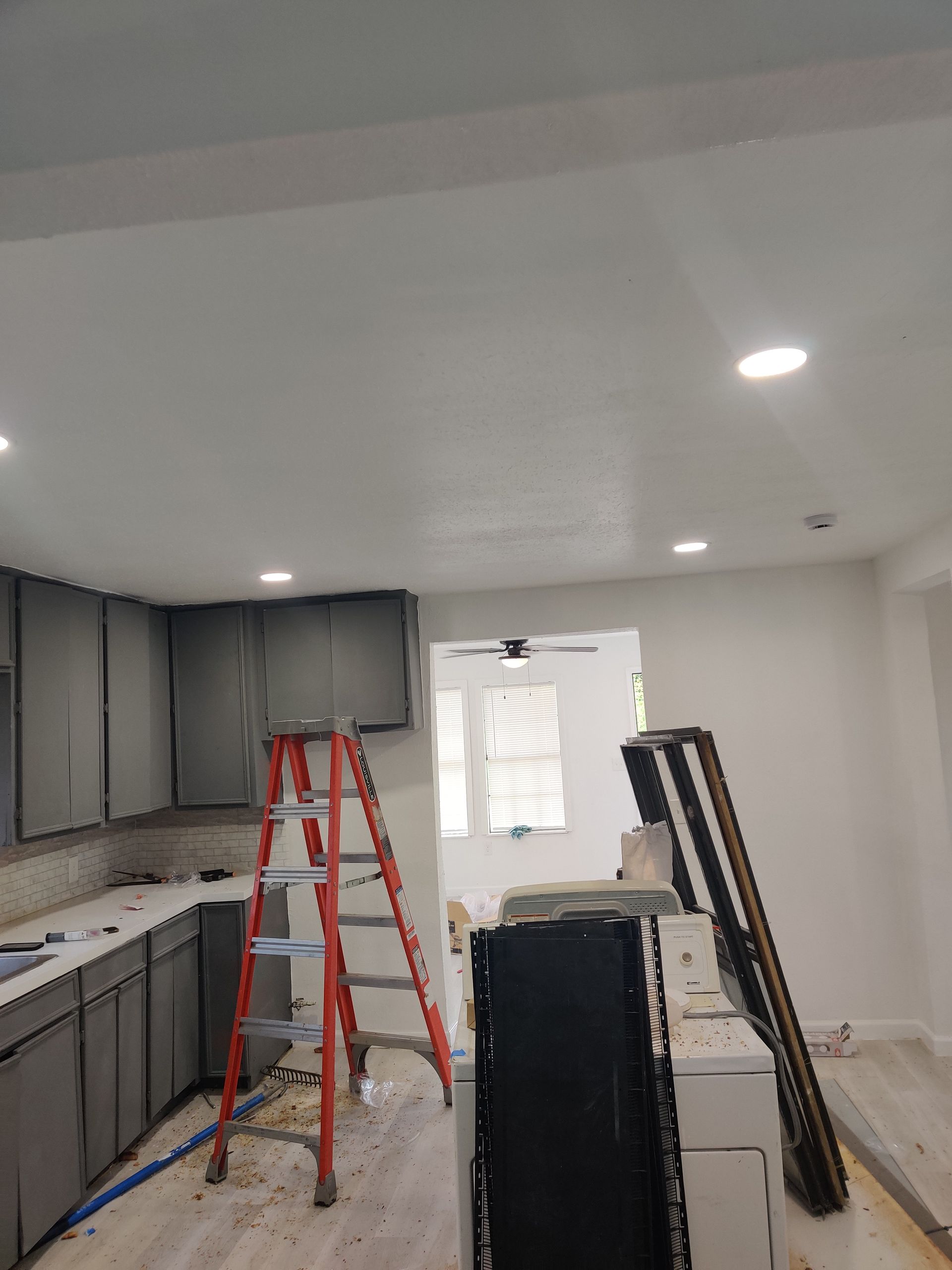 Kitchen renovation in progress: gray cabinets, orange ladder, white walls and ceiling, recessed lights.