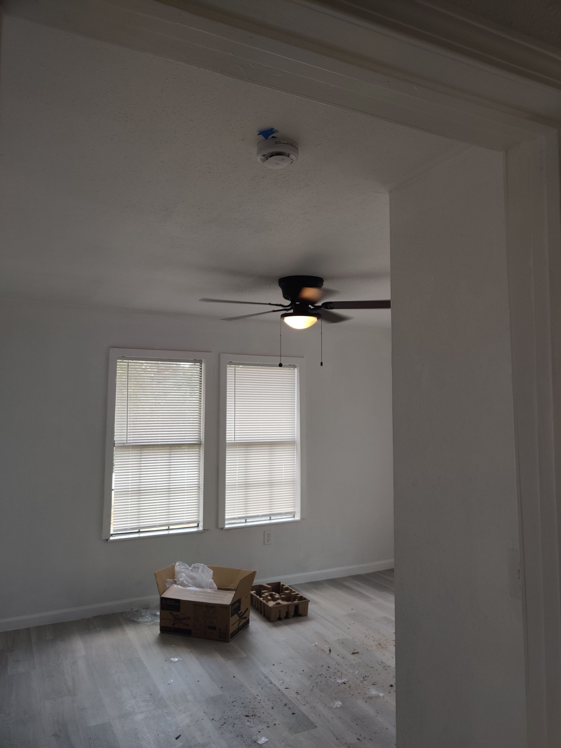 A room with two windows, a ceiling fan, and a smoke detector. Wooden crate and debris on the floor.