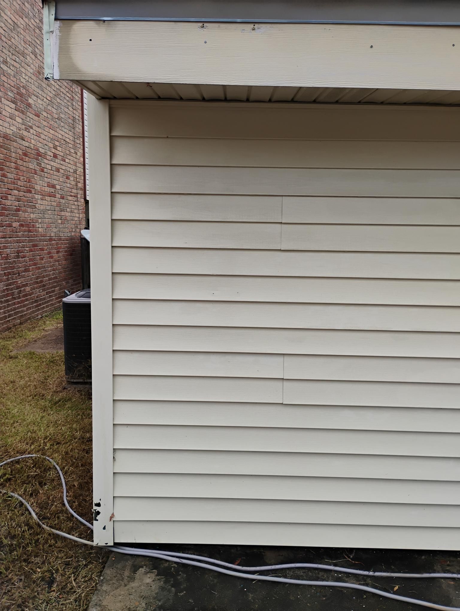 Beige siding on a building's side, possibly warped. Brick building and grassy area are visible.