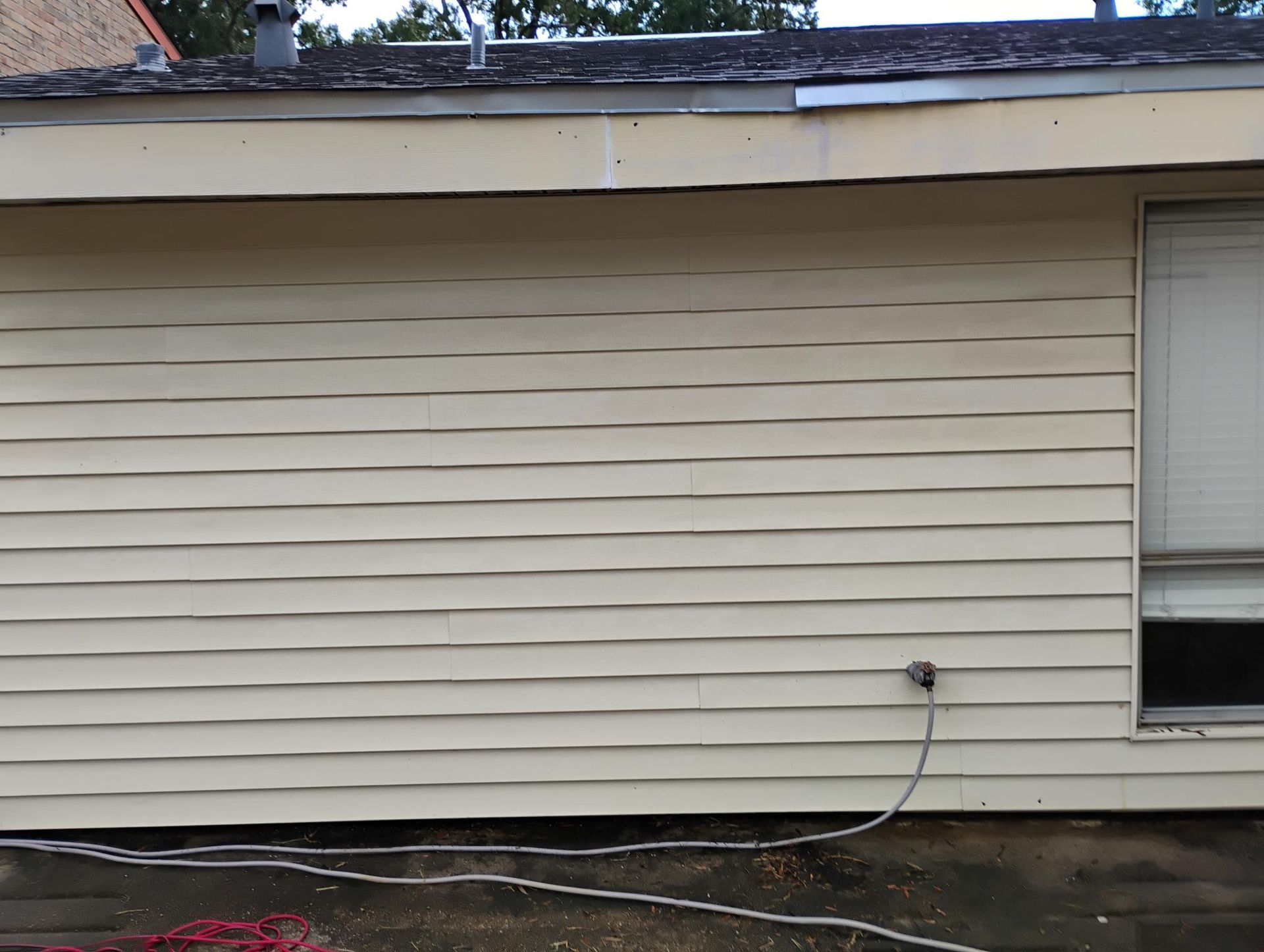 Exterior wall of a beige-sided building with a window and electrical outlet; a cable runs along the ground.