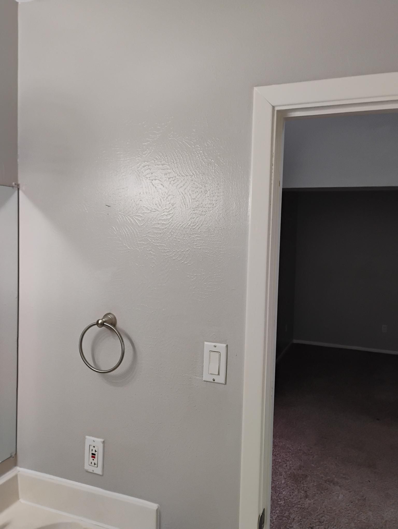 Gray bathroom wall with a towel ring, light switch, and doorway to a dark room.