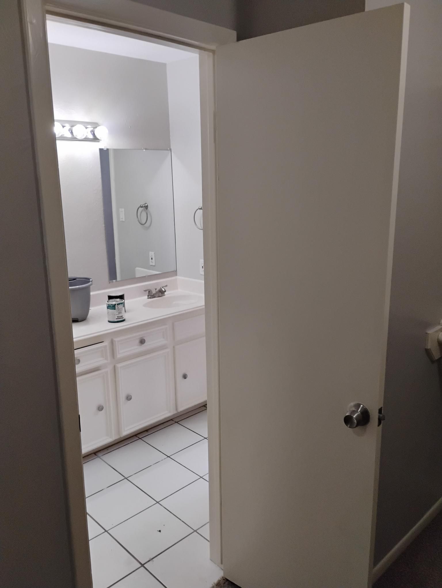 White door ajar, revealing a bathroom with a white vanity, mirror, and tiled floor.