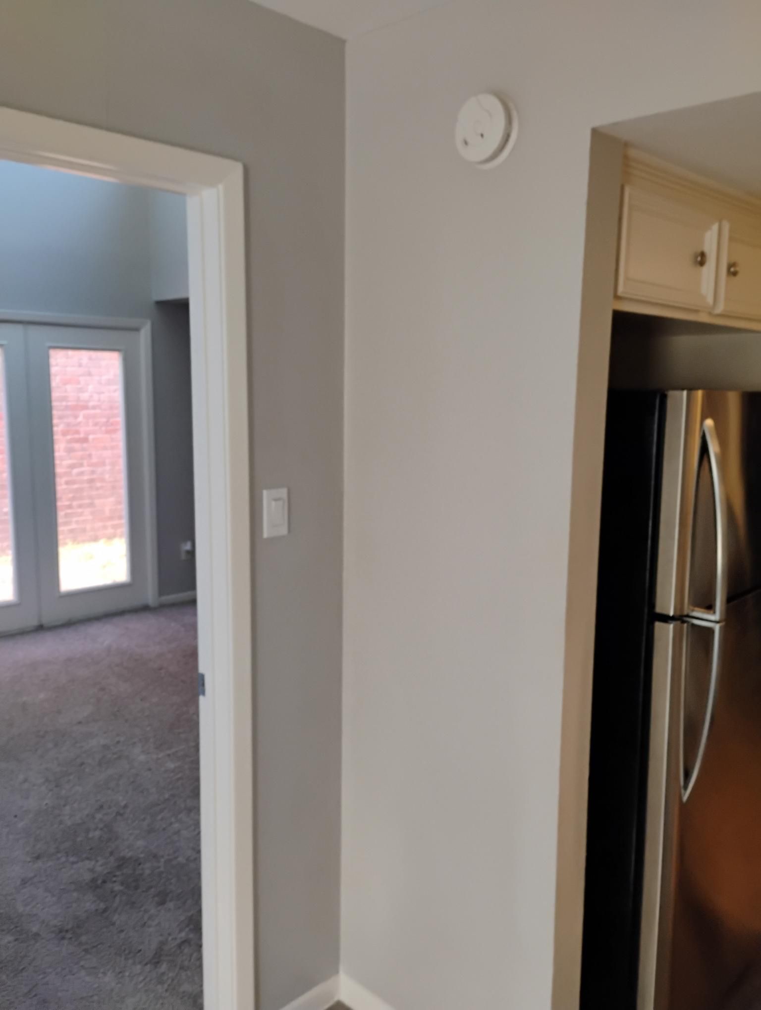 Gray interior wall with smoke detector, doorway to a room, and a refrigerator.