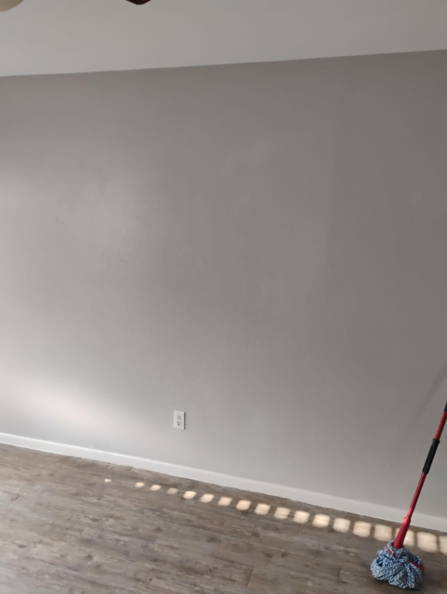 Gray wall with white trim, electrical outlet, and mop. Sunlight streams from the left.