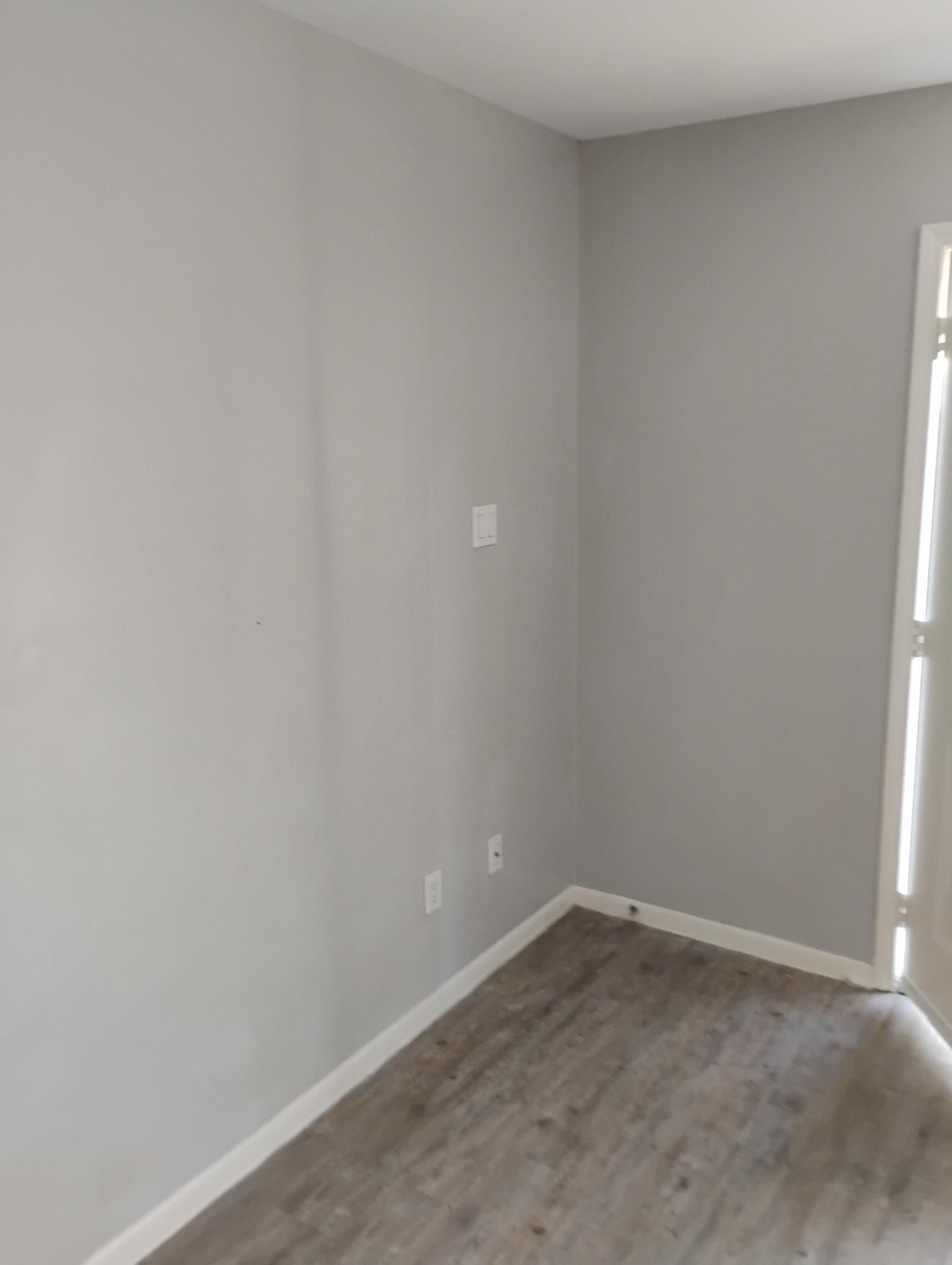 Empty corner room with gray walls, white trim, and wood-look floor. A door on the right.