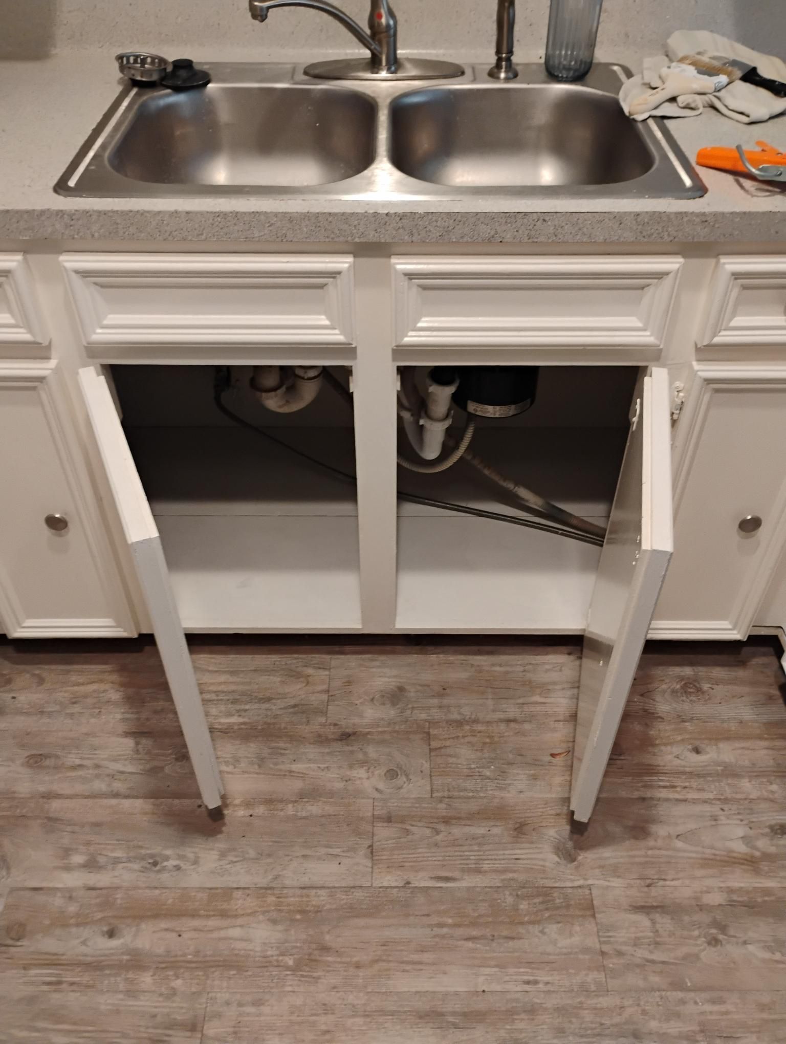 Kitchen cabinet under a sink with open doors, revealing plumbing and a garbage disposal.