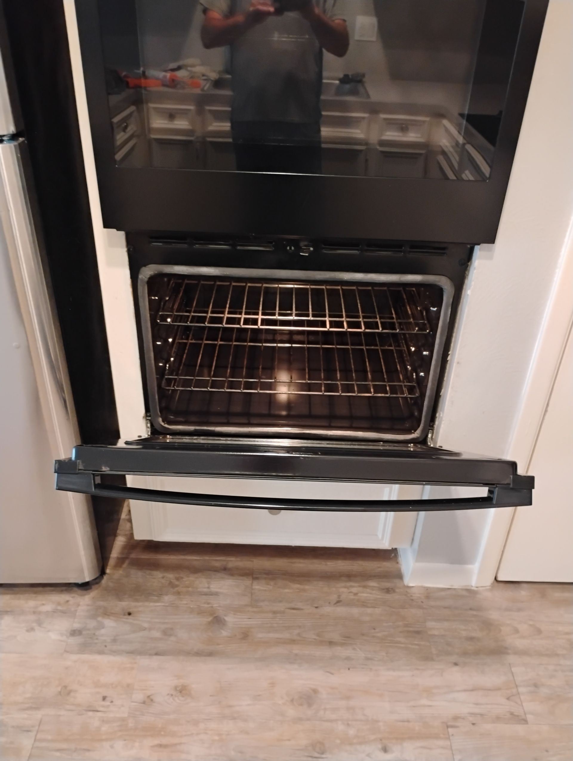 Open oven with racks inside, reflecting person taking photo. Black oven door, white trim, wooden floor.