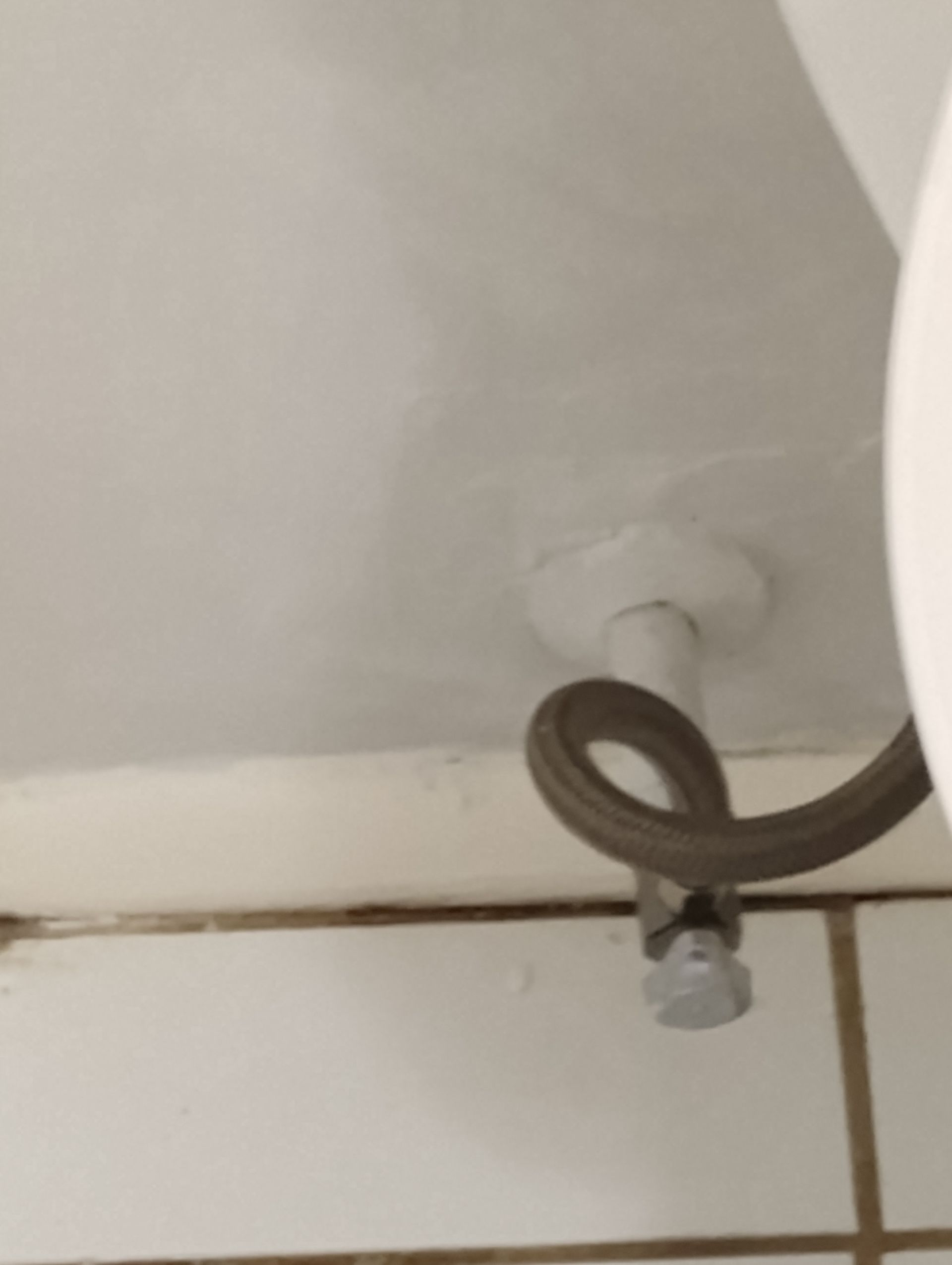 A white toilet water supply valve with a flexible hose connected, against a white wall and tiled surface.