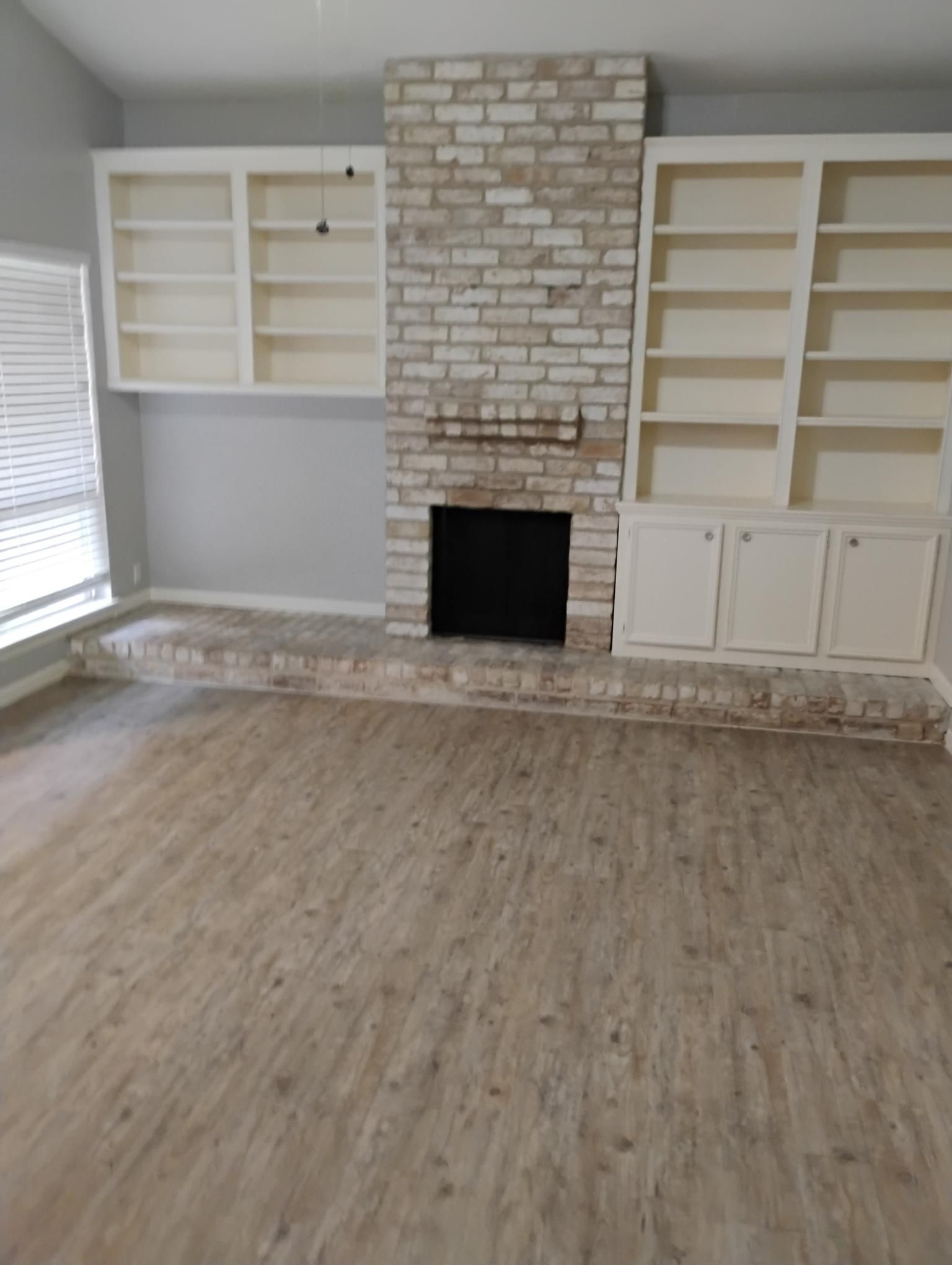 Room with a brick fireplace, built-in white bookshelves, and light-colored wood-look flooring.