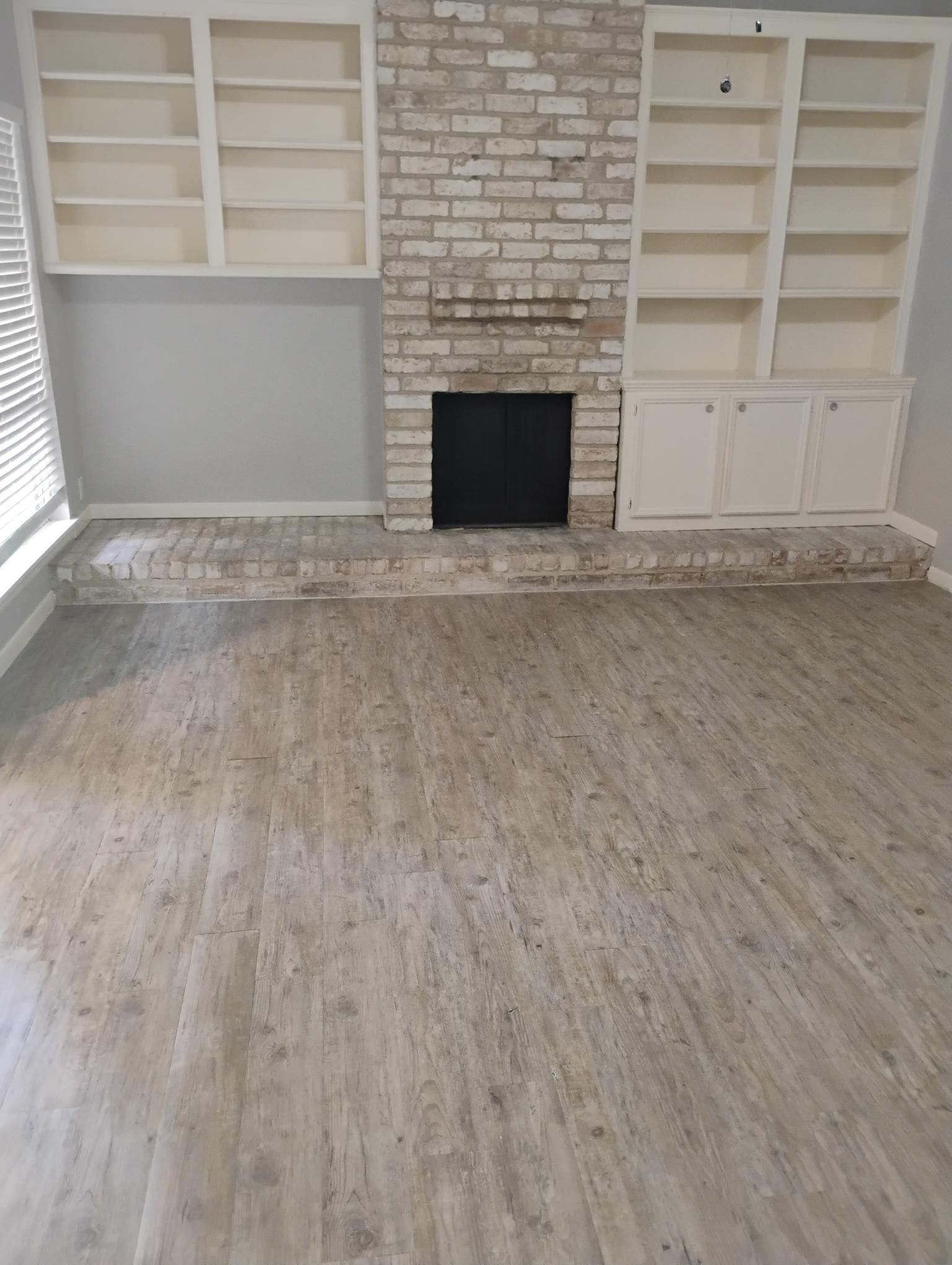 Living room with brick fireplace, built-in shelves, and wood-look flooring. Gray and white color scheme.