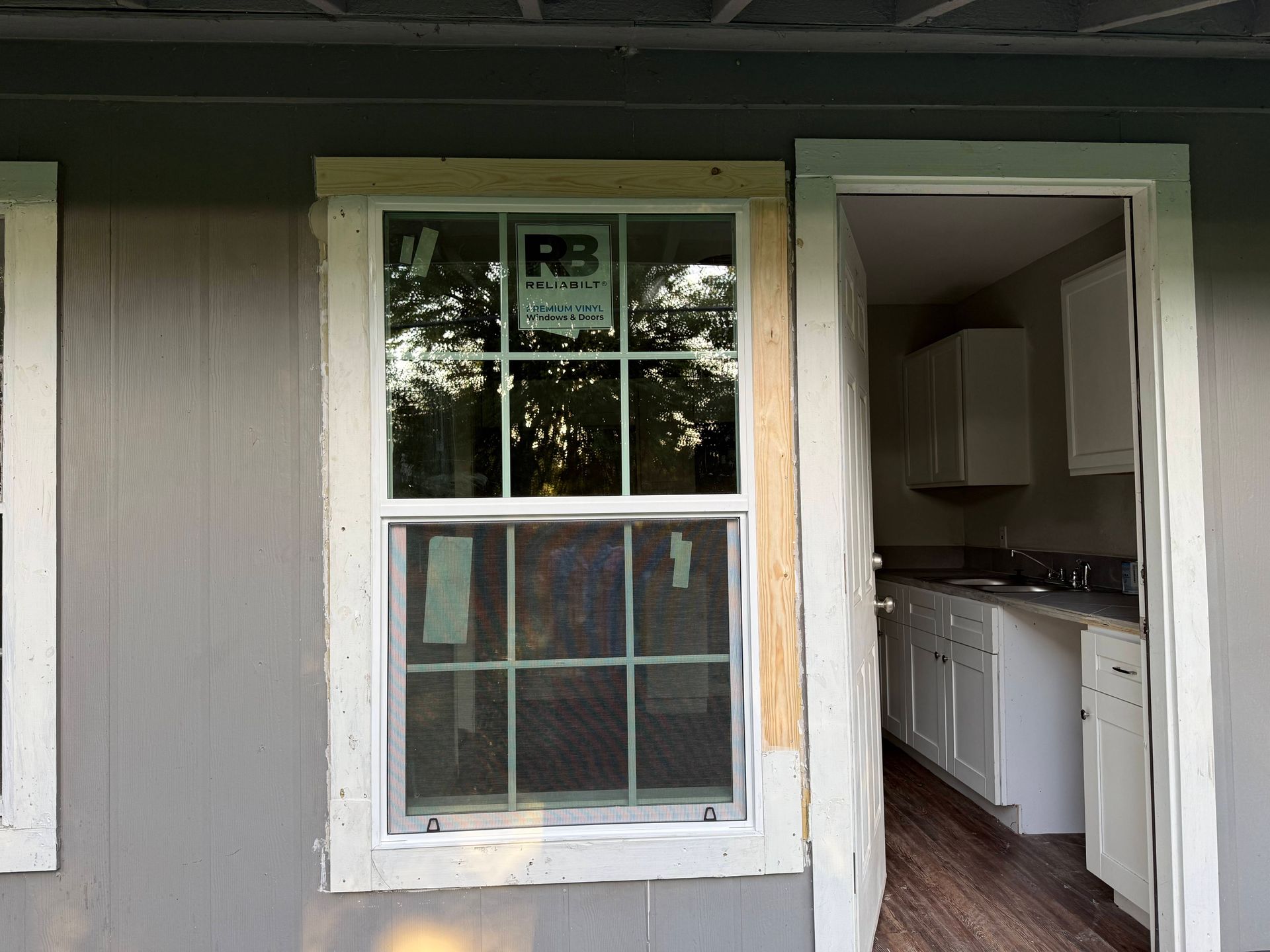 Exterior of building with window and doorway; grey siding, white trim, partial view of kitchen.