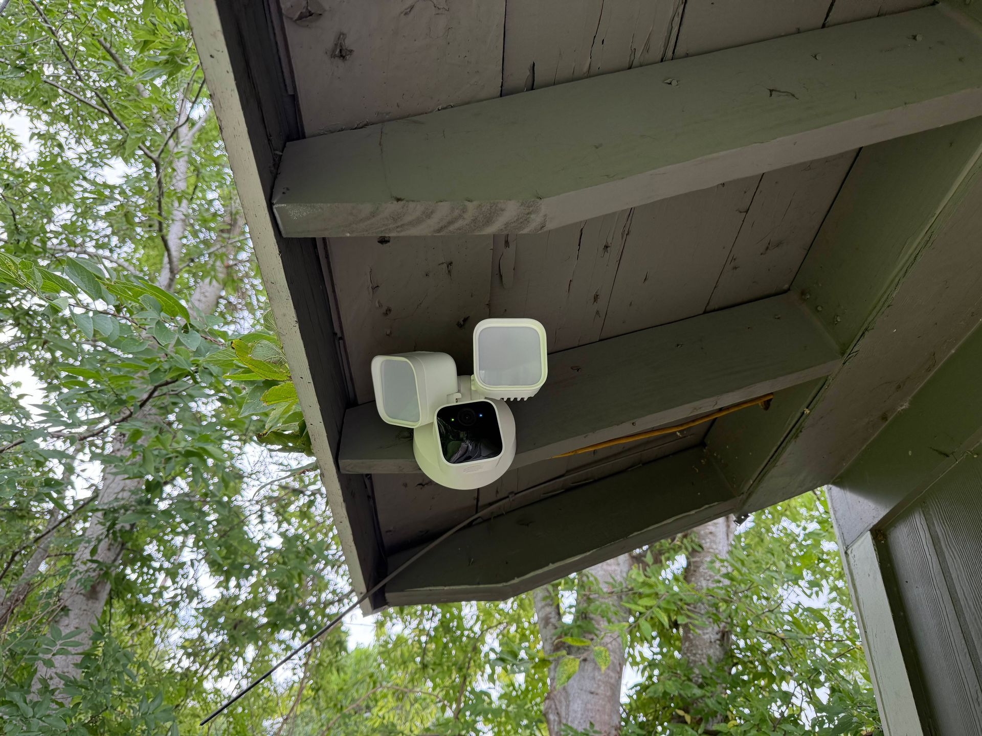 Security camera with three lights mounted under a green-painted wooden overhang, trees in the background.