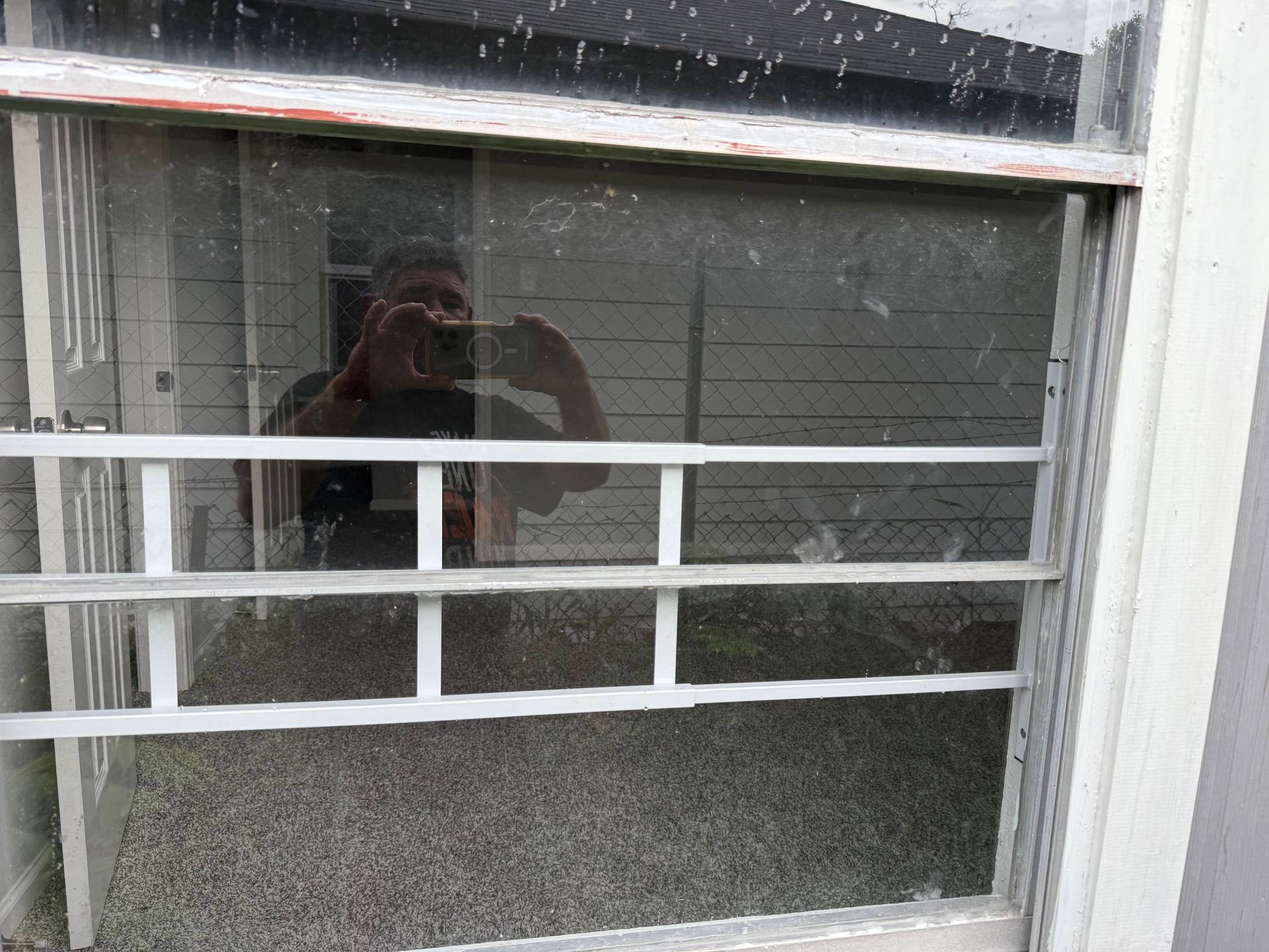 Person taking photo reflected in a dirty window with white bars. Backyard visible through the glass.