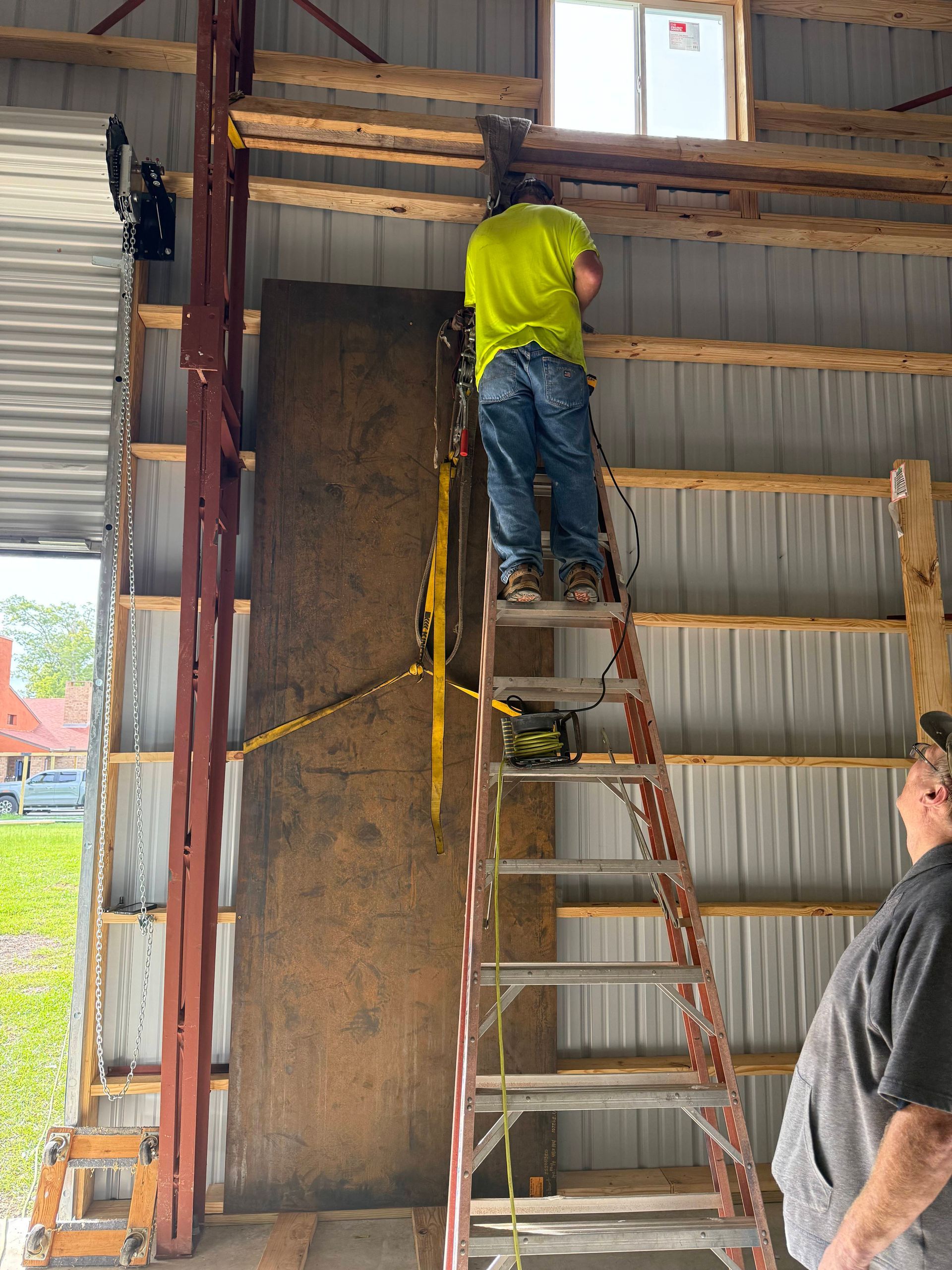 A person on a ladder working on a building's interior. A large plywood sheet is being positioned.