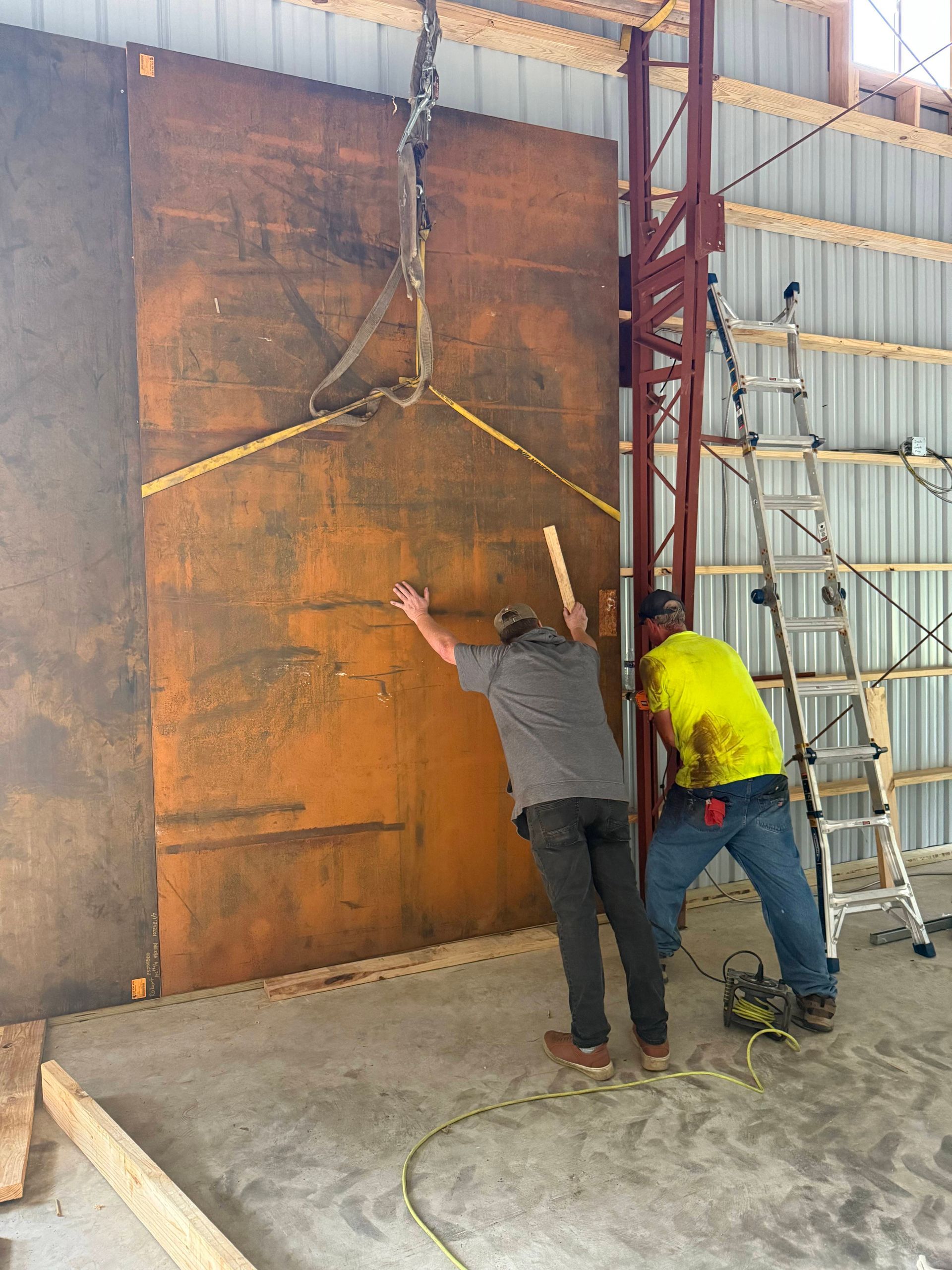 Two workers positioning a large, rusty metal sheet inside a building, with lifting equipment and a ladder nearby.