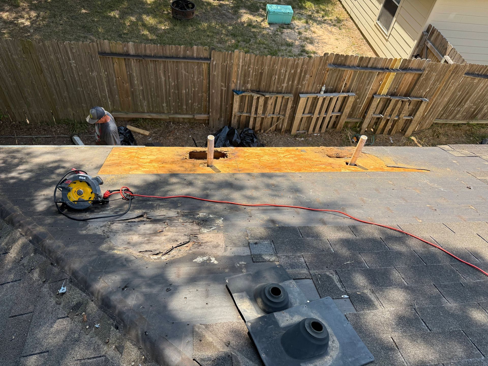 Roofer cutting plywood on a roof. A saw and power cord are visible. Brown fence, pallets, and yard in background.