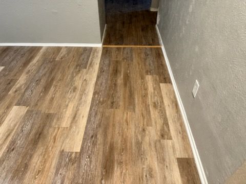 Wooden-look flooring in a room, meeting darker flooring in a hallway. White baseboards, light grey walls.