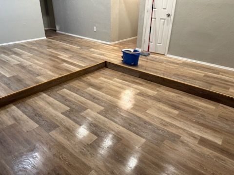 Newly installed laminate flooring with a brown border; a blue bucket and mop are visible.