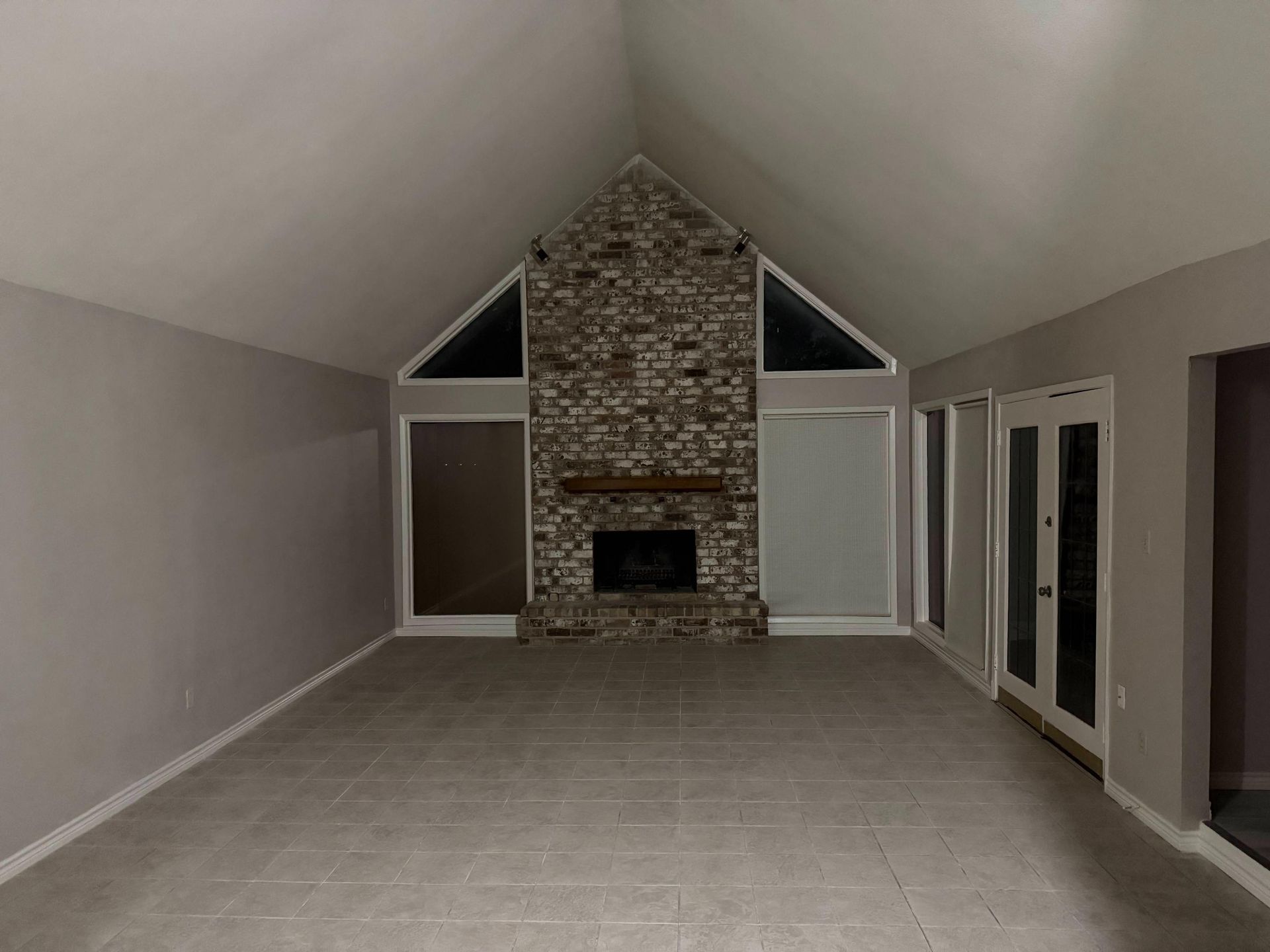 Empty living room with brick fireplace, high ceilings, large windows, and gray tile floor.